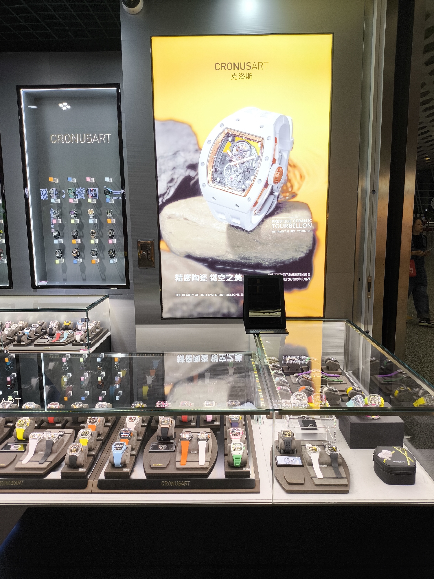 The image depicts a storefront display of watches, likely located in the Bao'an District, China. Two large illuminated signs feature the brand "CRONUSART," one showcasing a white ceramic tourbillon watch. Underneath, glass cases display a variety of watches on individual platforms, with prices listed. A black laptop is positioned on top of one of the display cases. An Asian male is walking outside, presumably looking into the store.