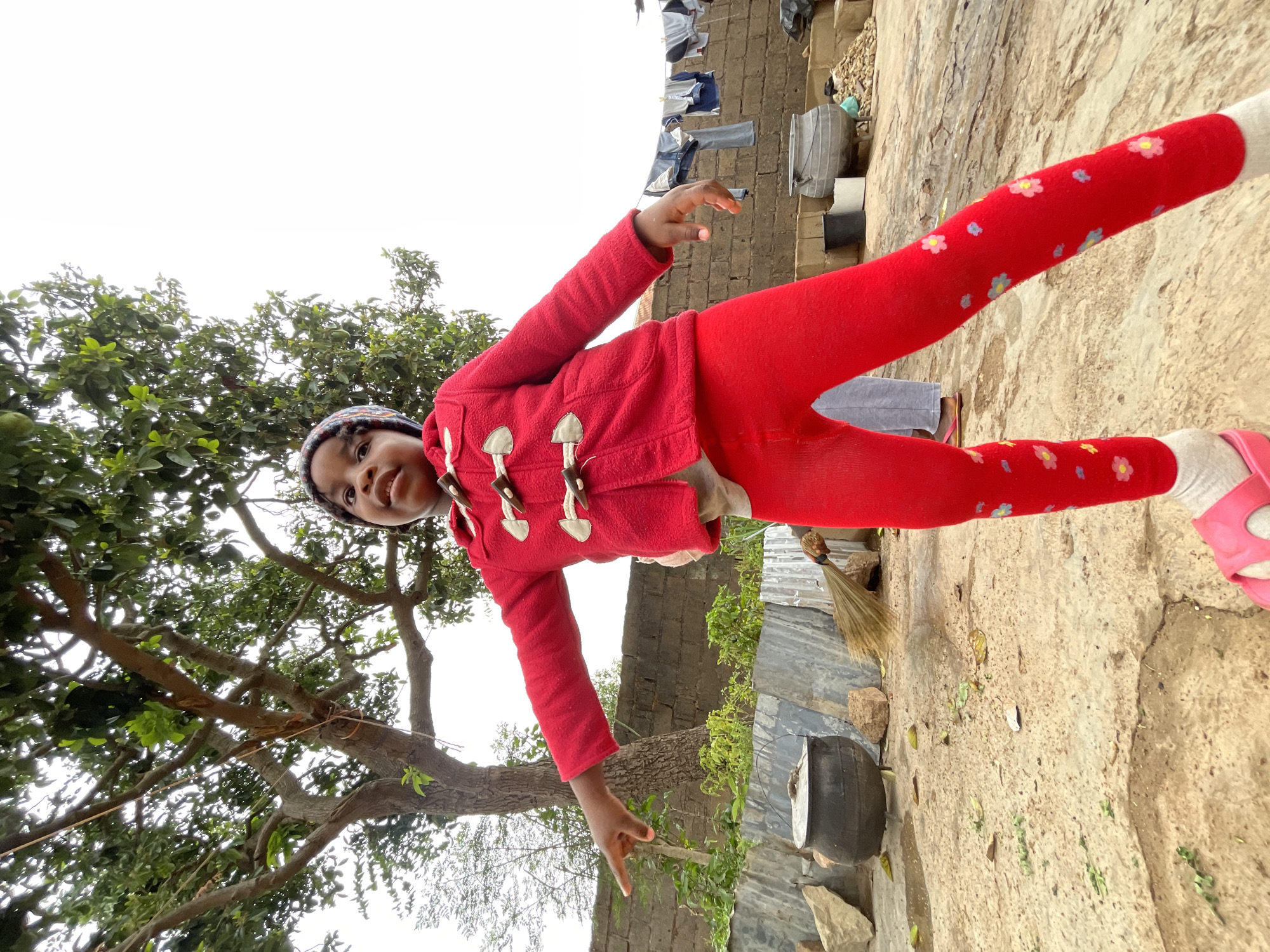 A young girl wearing a red jacket, red floral leggings, and a patterned beanie stands in an outdoor environment. She is gesturing with both hands. A tree with green leaves is positioned to her left. Behind her is a wall with a clothesline strung above. A broom leans against a metal sheet structure near a dark-colored pot on the ground. The ground appears to be a mix of cracked concrete and dirt. The photograph was likely taken in Jos, Nigeria, due to prior context.