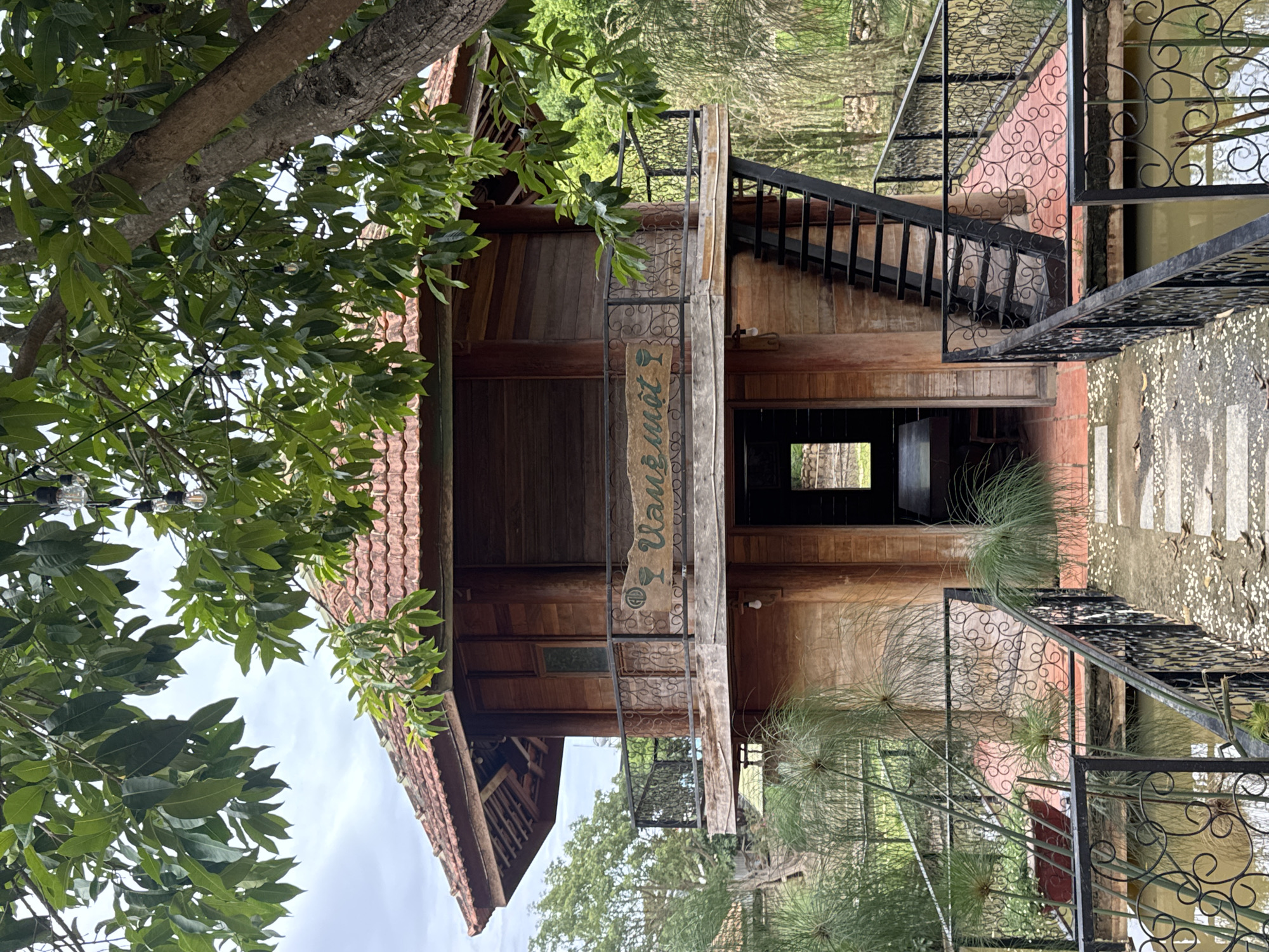 A wooden building with a red tile roof stands in Phú Thạnh, Vietnam, partially obscured by a tree with dense green foliage in the left foreground. The building features a set of metal stairs leading to an upper level and a sign with Vietnamese script hanging vertically. The structure is built above a body of water, with decorative metal railings along the walkway. The ground surrounding the building appears to be paved. The scene is outdoors, captured on an overcast day.
