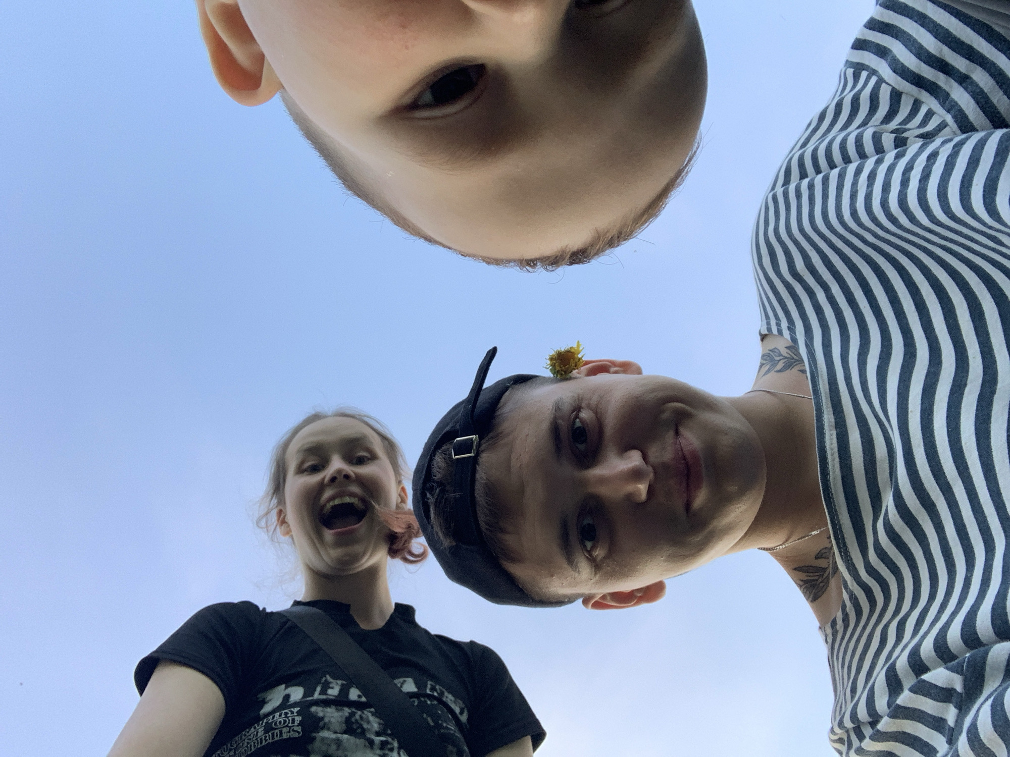 The image shows a low-angle view of three individuals. One is at the top, positioned almost directly above the camera, showing only a portion of their head. To the right is another person wearing a striped shirt, with a tattoo on their neck. To the left, a third person is wearing a black t-shirt and has an open mouth. The background is a clear blue sky. The image doesn’t contain any information about the city Фоки, Russia.