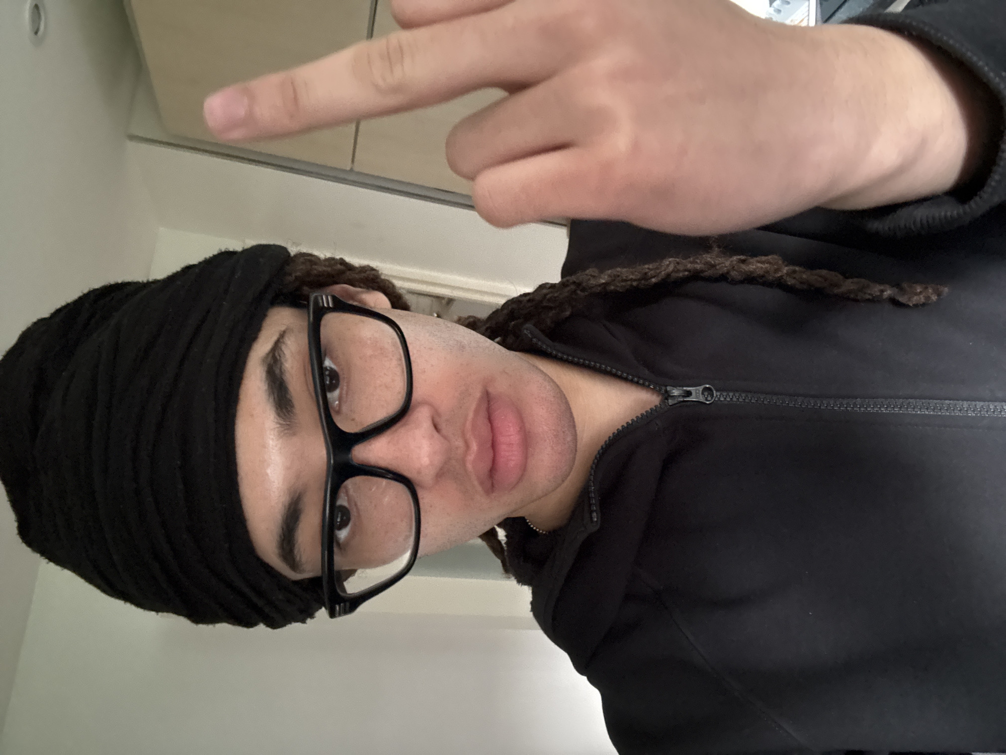 A person with dark hair and glasses, wearing a black head covering and a zippered black jacket, poses for a selfie. The person's right hand is extended, displaying the middle finger. The background includes a white wall and a light-colored cabinet. The location is Les Ulis, France.