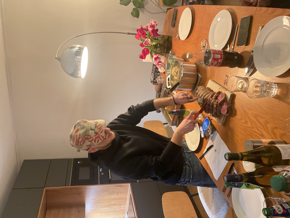 A person in a patterned cap carves a roast on a wooden cutting board at a dining table laden with plates, drinks, and a pot of food. Tulips bloom in a vase beside the feast. Bottles of wine and other beverages are scattered near empty plates. A modern, arched floor lamp illuminates the scene. In the background are dark kitchen cabinets and appliances.