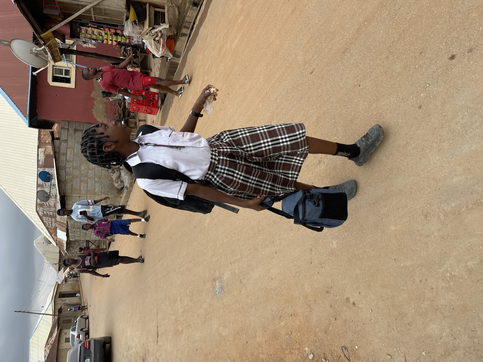 A young woman in a school uniform walks along a dusty street in Karmajiji, Nigeria. She wears a white shirt, a plaid skirt, black socks, and gray athletic shoes. A black backpack is slung over one shoulder, and a blue bag is in her hand. She appears to be holding a food item in a plastic bag. Other people walk along the street. Buildings with corrugated metal roofs line the street. Cars are parked further down the road. A storefront with snacks and beverages displayed is in the background. A satellite dish is affixed to one of the buildings.