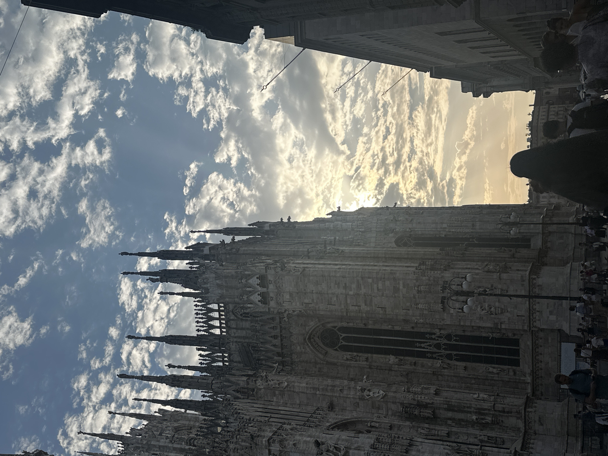 An eye-level, medium shot captures the facade of a Gothic cathedral on the left, towering over a busy street in what appears to be an urban environment. The cathedral's intricate stone architecture, adorned with spires and detailed carvings, is partially silhouetted against a bright, cloudy sky. The sky is a vibrant blue, with scattered, fluffy white clouds illuminated by the warm glow of the setting sun, casting long shadows across the scene.
The street in the foreground is bustling with activity. Several people are walking, some with backpacks, suggesting they might be tourists. A man in the lower left is walking towards the camera, looking down at his phone. Street lamps with multiple globes are visible along the side of the cathedral. To the right of the cathedral, a modern building rises, its upper floors partially obscured by the frame. The overall mood is serene yet lively, with the grandeur of the historical architecture contrasting with the everyday life unfolding on the street.