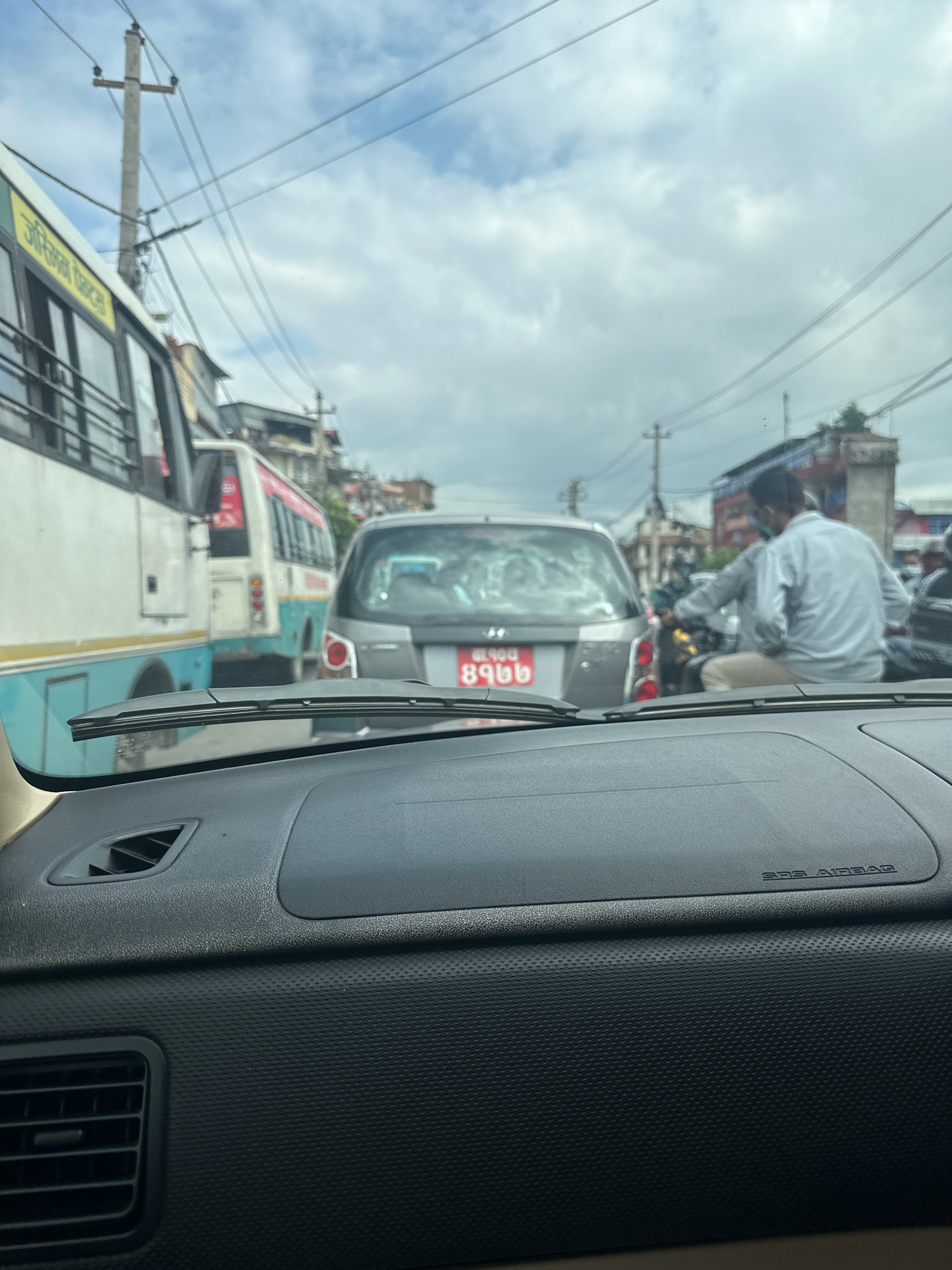 The image shows a view from inside a vehicle, looking forward at a traffic jam on a city street, likely in Kathmandu Metropolitan City, Nepal. The foreground consists of a car's dashboard, with a windshield wiper visible at the bottom. Beyond the windshield, a silver car is stopped, displaying a red license plate with Nepali script. To the left of the silver car, a white and blue bus is partially visible, also stopped. Further in the background, another bus and buildings line the street, and power lines are strung across the sky. A man on a motorcycle is in the lane next to the silver car, and another person is on a scooter behind him. The sky is cloudy.