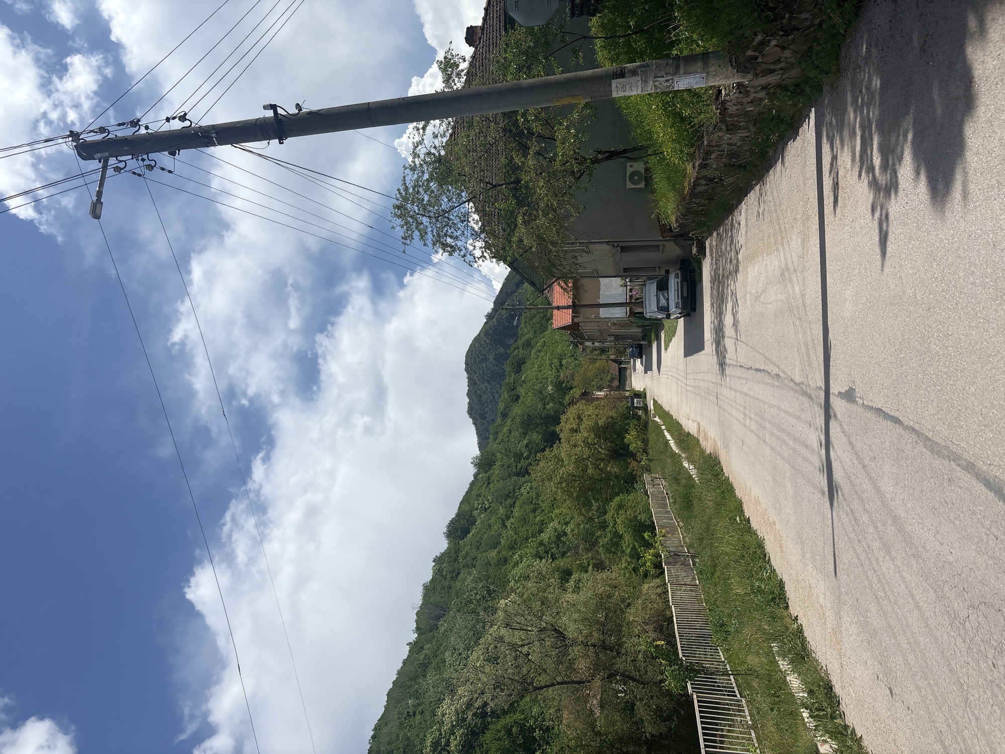 The image captures a narrow asphalt road in a neighborhood of Unknown, Unknown. The road is bordered on one side by a lush green hillside covered with trees, and on the other side by houses and buildings. A tall utility pole with wires runs along the side of the road. The sky is partially cloudy, with patches of blue visible. A silver van and other parked cars are visible along the road. A long set of stairs leading up the hillside is visible on the right side of the road. The lighting suggests it is daytime.