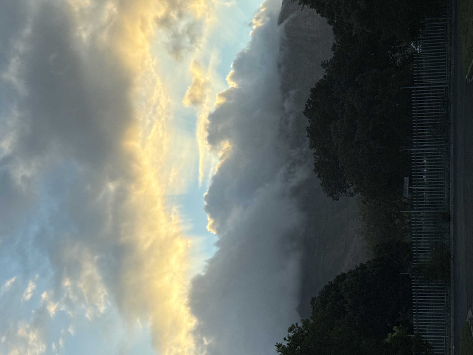 The image presents a view of clouds over what appears to be a mountainside in Cape Town, South Africa. The sky features a mix of grey and gold-tinged clouds, with glimpses of blue sky. The mountainside is obscured by a bank of heavy cloud or mist. Trees are visible along the base of the mountain. A metal fence and a street sign are visible along the right side. The street sign has the inscription "Highlands Lane" visible.