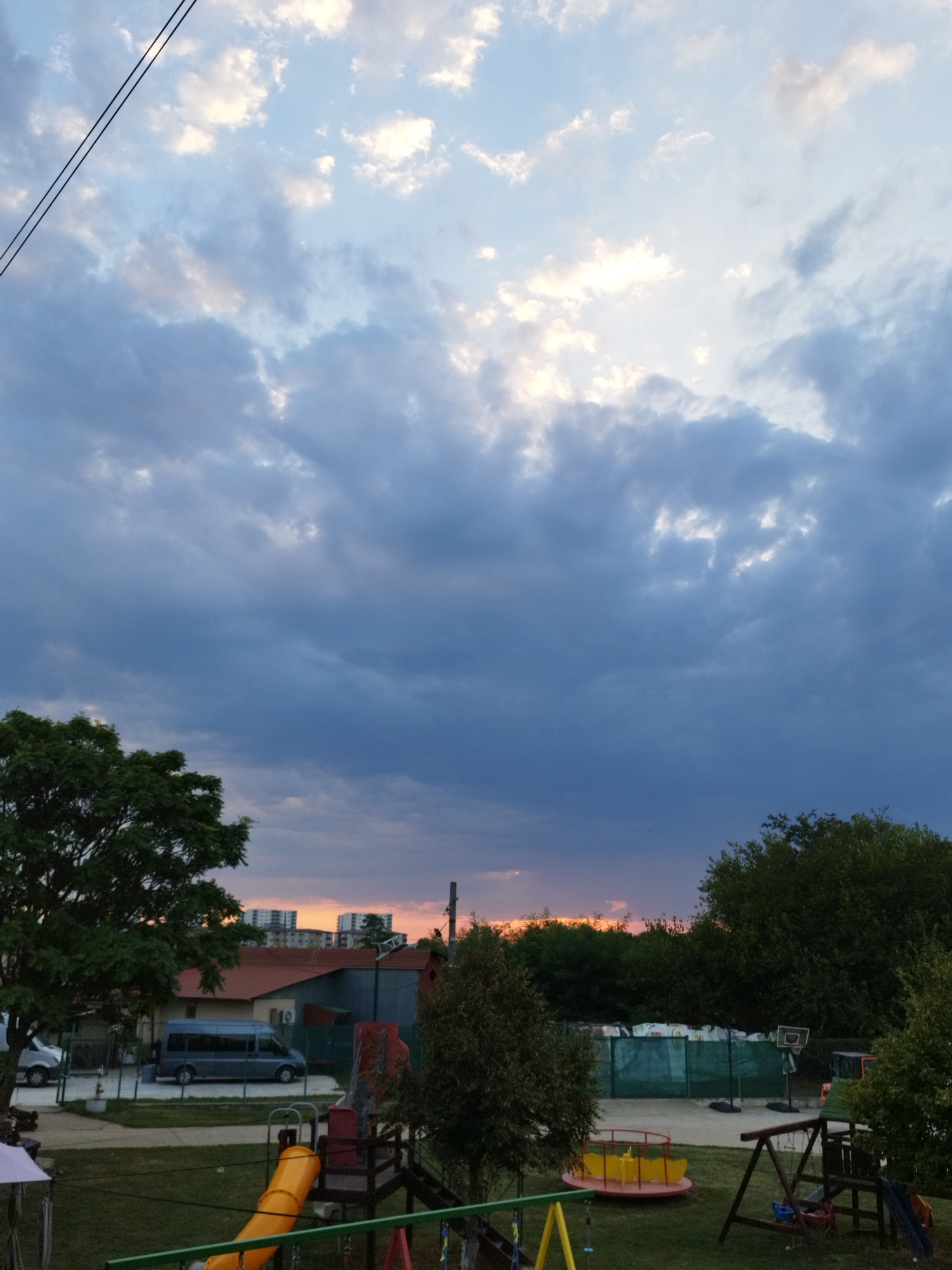 The image depicts a suburban scene at dusk, likely in Bucharest, Romania. The sky is filled with dramatic clouds, with patches of the setting sun visible through them, casting an orange glow on the horizon. In the foreground, a children's playground is visible, featuring a yellow slide, a merry-go-round, and a swing set. Adjacent to the playground is a grassy area. Further back, a street is lined with buildings and trees. A blue minivan is parked on the street, and a larger building with multiple windows is visible in the background. The overall impression is one of a peaceful, end-of-day setting in a residential area.