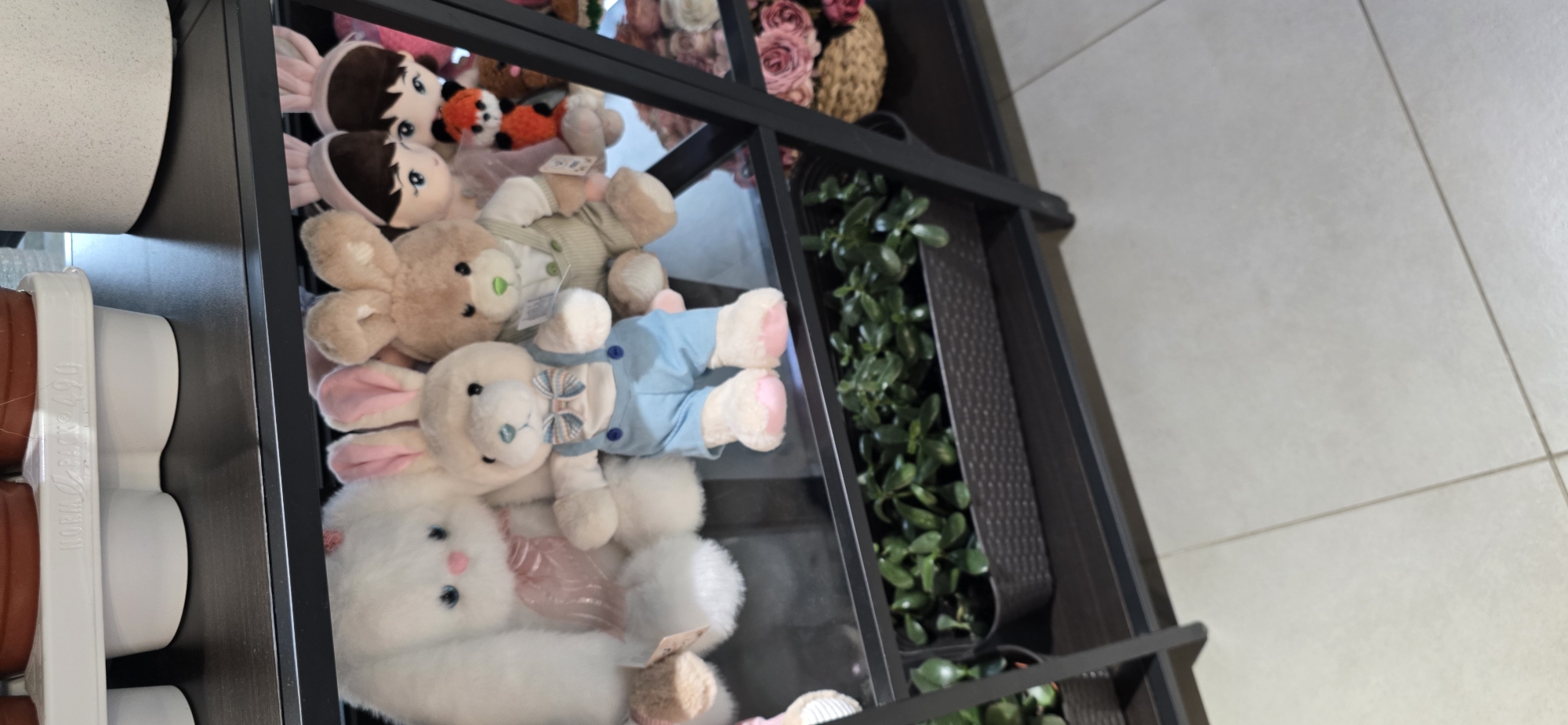 In a retail setting, possibly a shop in Русская Деревня-2 Кп, Russia, a black metal shelving unit holds various items. The top shelf displays white planters and a large white stuffed bunny with a pink ribbon. Below that, a second shelf is filled with several stuffed animals, including a white bunny in blue overalls and a bow tie, a brown bunny in a vest and shirt, and other assorted plush toys. The bottom shelf contains a dark brown woven planter filled with lush green plants. A woven basket filled with pink roses is visible on the right side of the bottom shelf. The overall environment appears to be well-lit, suggesting an indoor retail space.