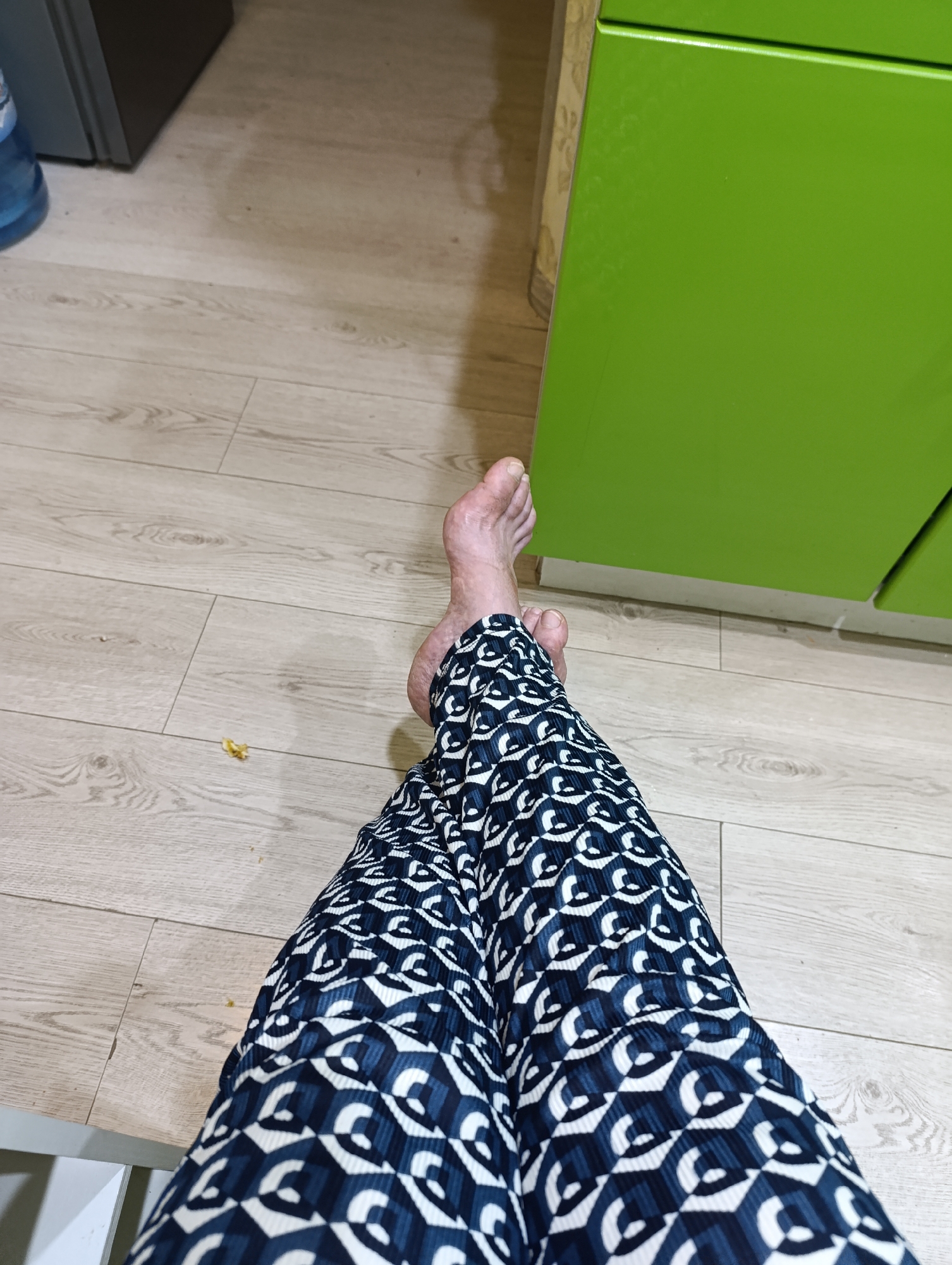The image shows a close-up view of a person's legs and feet wearing patterned pants, sitting on a wooden floor. The pants have a geometric pattern of dark blue and white shapes. One bare foot is visible, resting on the floor. To the right of the legs, a section of a bright green cabinet or furniture is visible. In the background, part of a grey appliance and a blue water dispenser can be seen. The floor is made of light-colored wood planks. There is no indication of the location being in Tugulym, Russia, or any specific city. The scene appears to be indoors, possibly in a kitchen or living area.