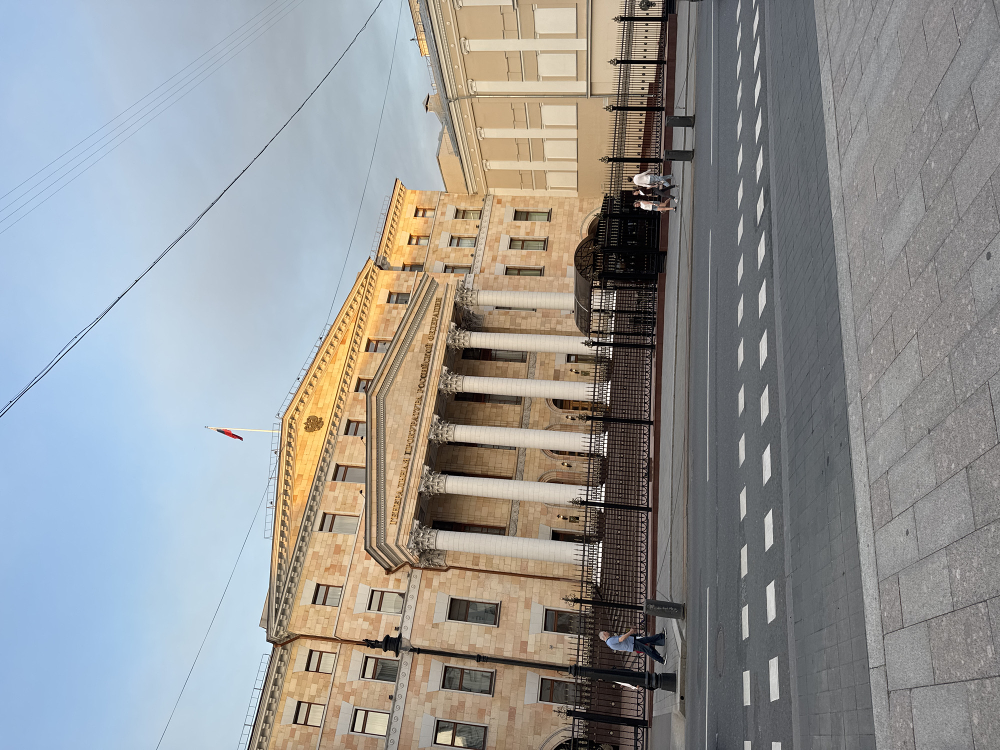 The image shows the General Prosecutor's Office of the Russian Federation building in Moscow, Russia. The building is made of light brown stone and features a grand entrance with large white columns. A Russian flag is visible on top of the building. Several people are walking on the sidewalk and crossing the street in front of the building. The sky is a light blue with some clouds. There is a black metal fence in front of the building and a street with a pedestrian crossing. The lighting suggests it is either early morning or late afternoon.