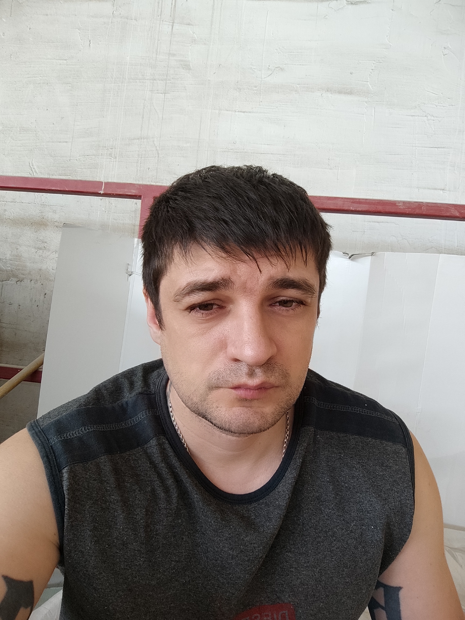 A man is shown in a close-up, head-and-shoulders selfie. He is facing the camera directly with a neutral expression. He has short, dark hair and is wearing a dark gray sleeveless shirt with black detailing across the shoulders. A silver chain necklace is visible around his neck. The background appears to be an indoor setting with a textured white wall and a red bar running horizontally across the top of the image. To the left of the man, a portion of a wooden pole or beam is visible. There are also white panels or sheets in the background, suggesting some sort of construction or renovation site. The lighting is somewhat harsh, casting subtle shadows on the man's face.