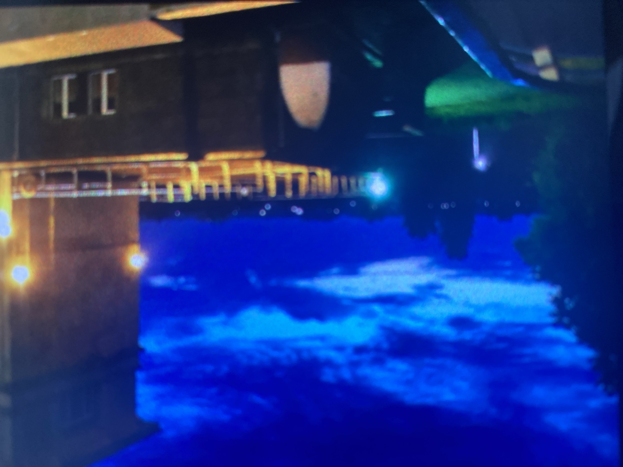 An upside-down night scene. The top half features a dark building with lit windows and what appears to be a bridge or walkway with lights along it. The bottom half depicts a body of water with a reflection of the building and sky. The sky is dark blue, possibly indicating night.
Although I do not have enough information to identify the location as Saint Petersburg, Russia, I can note the architecture and the presence of canals are consistent with the city.