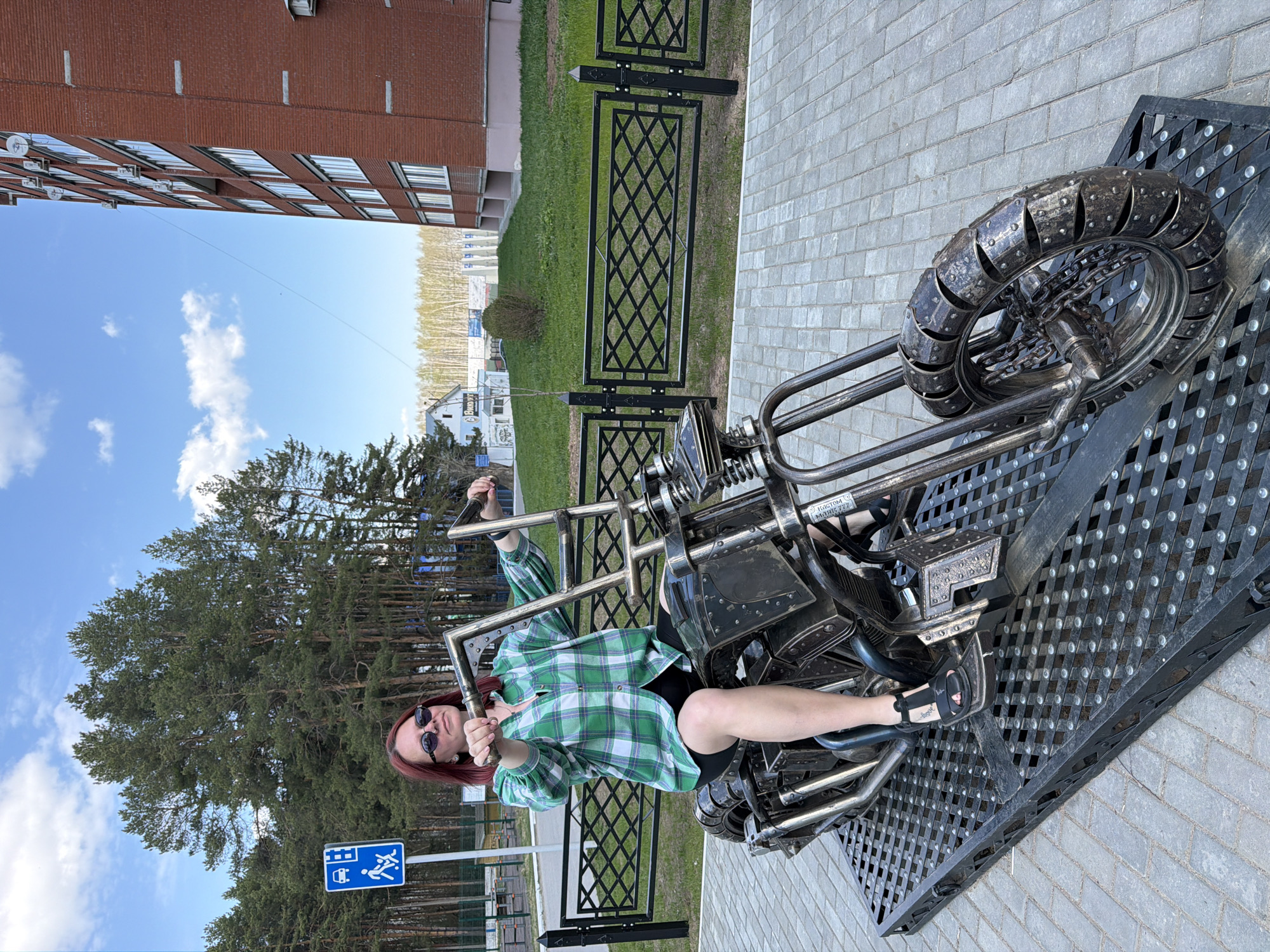 A photograph taken in Chaikovsky, Russia shows a woman sitting on a steel motorcycle sculpture. She has red hair, wears sunglasses, a green and white plaid shirt, and black shorts. She is making a hand gesture. The motorcycle is detailed with metal plates and a studded tire. The sculpture sits on a platform of metal grating, which is placed on a brick walkway. A black metal fence borders a grassy area with trees behind it. In the background, there is a red brick building under a partly cloudy blue sky. A blue sign is visible near the woman.