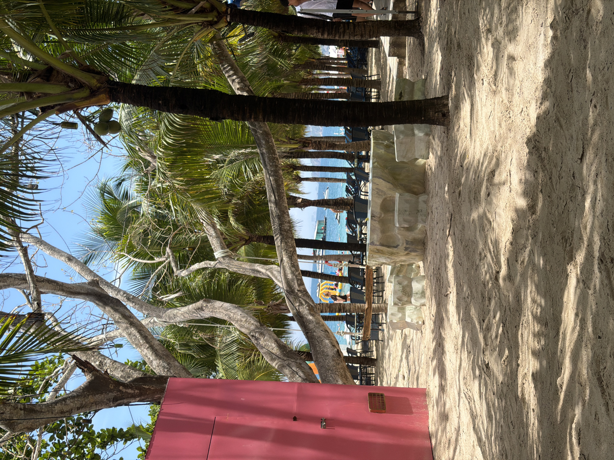 The image depicts a beach scene in Nam Nha Trang Ward, Vietnam. A row of palm trees lines the beach, with chairs and tables set up underneath them. The foreground is dominated by sand and a red structure. In the background, the ocean is visible with a few people and boats. The scene is bright with sunlight.