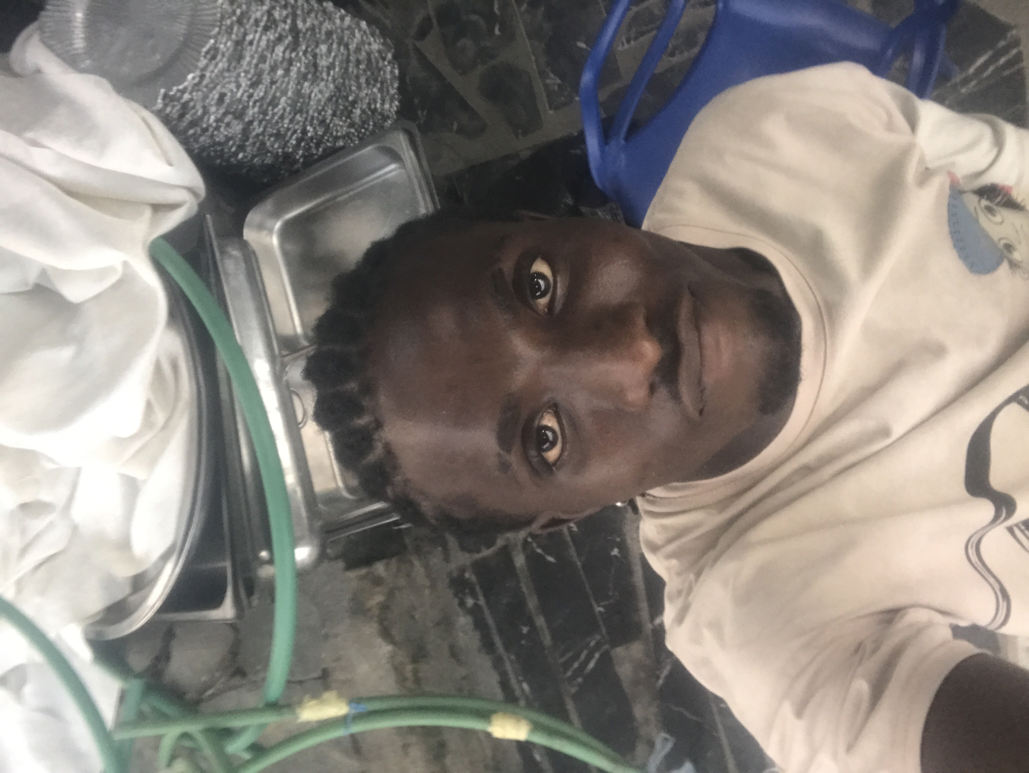 The image, oriented sideways, features a dark-skinned individual with short, dark hair or dreadlocks, looking directly into the camera. The person occupies the central and right portions of the frame and wears a light-colored, possibly off-white, t-shirt or sweatshirt. A graphic design, resembling a stylized face with large eyes, is partially visible on the right side of their chest.
To the upper left of the person's head, a blue plastic chair is visible. Further to the left and top of the frame, several metallic objects are arranged. These include a large, round metallic basin partially obscured by a white cloth, and multiple rectangular metal trays or containers, possibly for food service. A textured, silvery object, resembling a scouring pad or metal brush, is also visible among these items.
In the foreground, a prominent bright green hose or tube snakes across the lower left and central parts of the frame, marked with segments of yellow tape. The background consists of a dark, possibly tiled or concrete floor, indicating an indoor or semi-enclosed functional environment such as a kitchen, catering space, or storage area. The lighting is ambient. The person's expression is neutral, suggesting a selfie. No discernible text is visible in the image.
