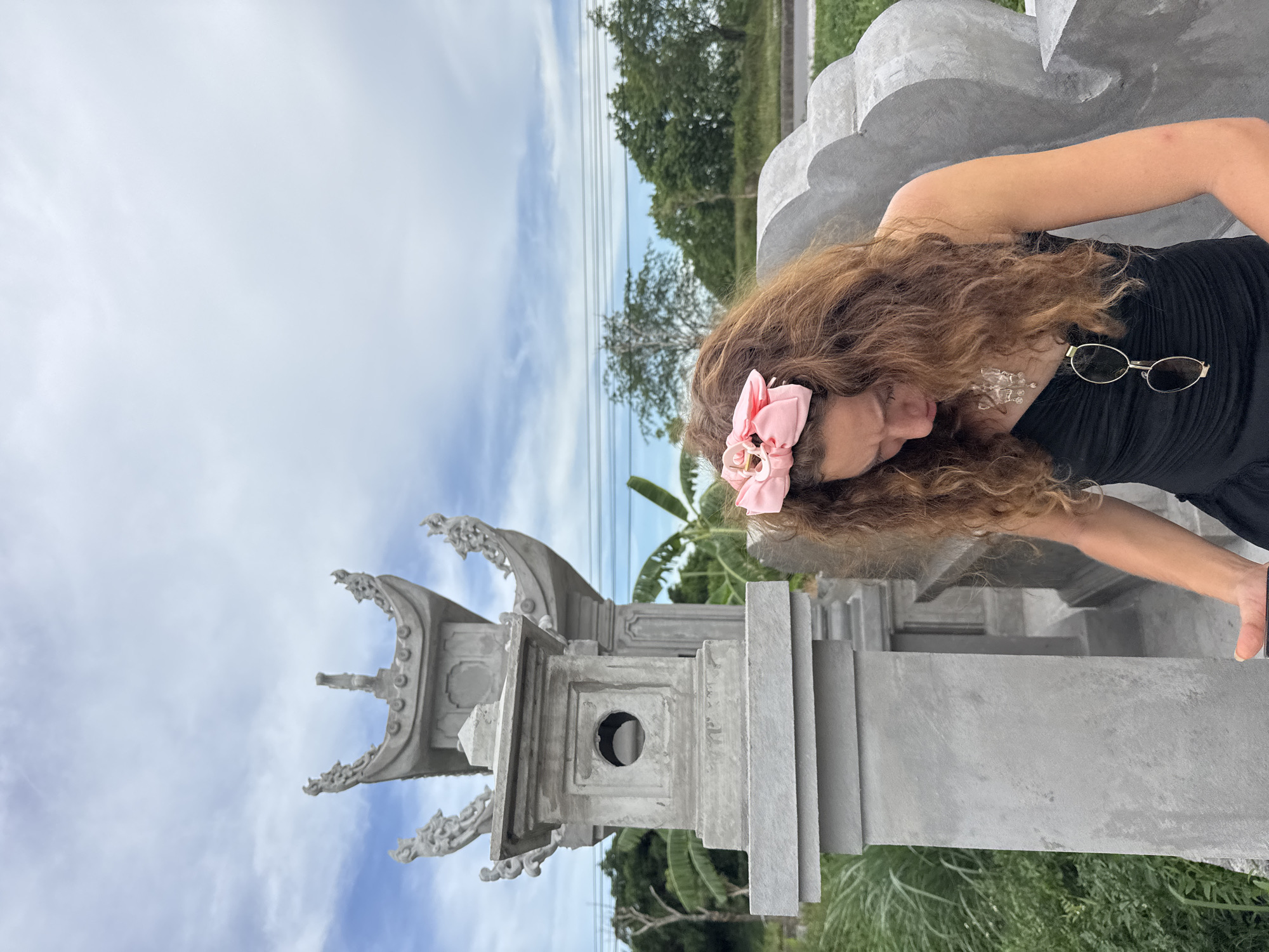 A young woman with curly brown hair, wearing a black dress and a pink bow in her hair, stands in front of a stone structure with ornate roofing. Sunglasses are hanging from her dress. She is looking down, with a necklace visible around her neck. The stone structure appears to be a monument or a temple, with intricate carvings and architectural details. In the background, lush green vegetation and trees can be seen under a cloudy sky with visible power lines. The scene appears to be outdoors in a location resembling Hoa Lư Ward, Vietnam, known for its historical and natural beauty.