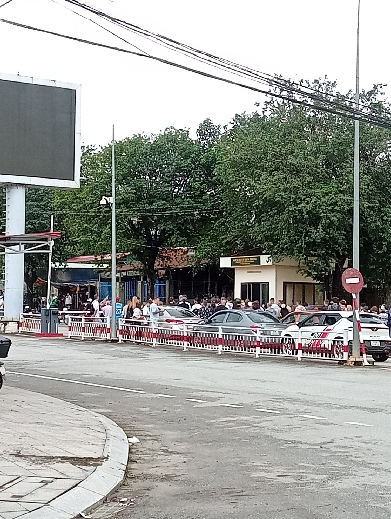 The image depicts a street scene in Bo Y, Vietnam, with a crowd of people gathered near a checkpoint. A white building with signage, possibly a border or security post, is visible in the background, surrounded by trees. Several cars are parked or stopped in front of a white and red striped barrier that separates the road from the crowd. People are standing and interacting, some appearing to be waiting. A security camera is mounted on a pole near the building. The street in the foreground is paved and shows signs of wear. Electrical wires are strung across the top of the frame.