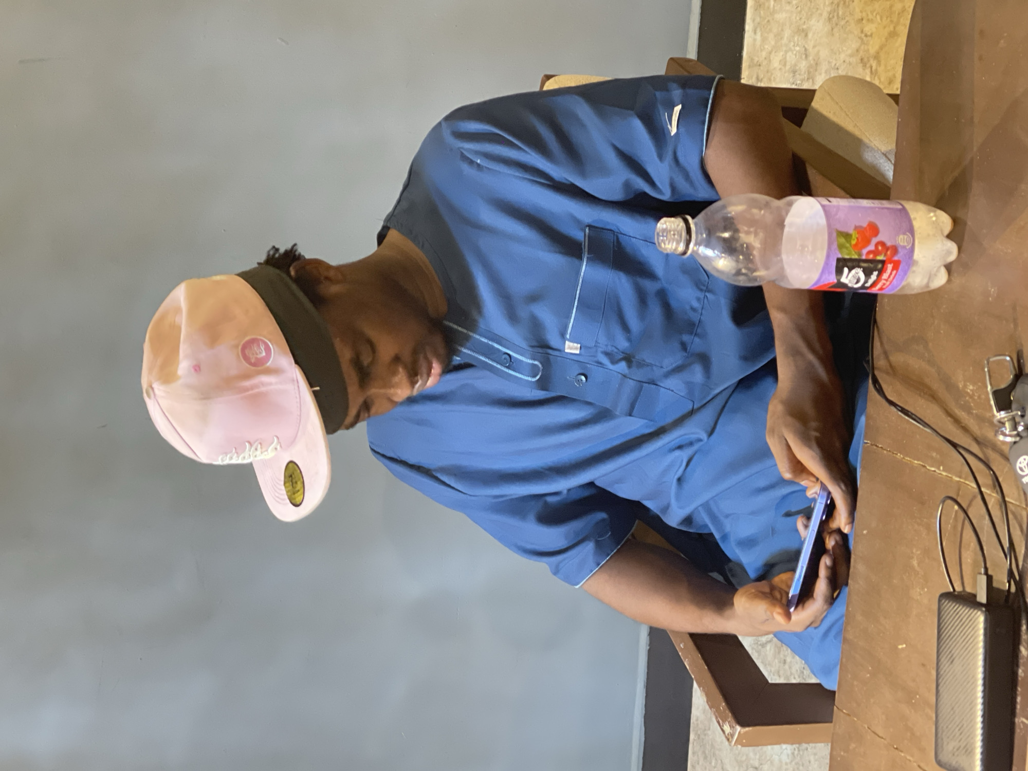 A young Black male is sitting on a wooden chair in front of a wooden table. He is wearing a blue traditional African garment and a pink baseball cap. He is looking down at his phone and holding it in his hands. On the table in front of him, there is a bottle of drink, a portable charger, and a black power bank. The background is a plain grey wall. The scene appears to be indoors, possibly in a casual setting within Abuja, Nigeria.
