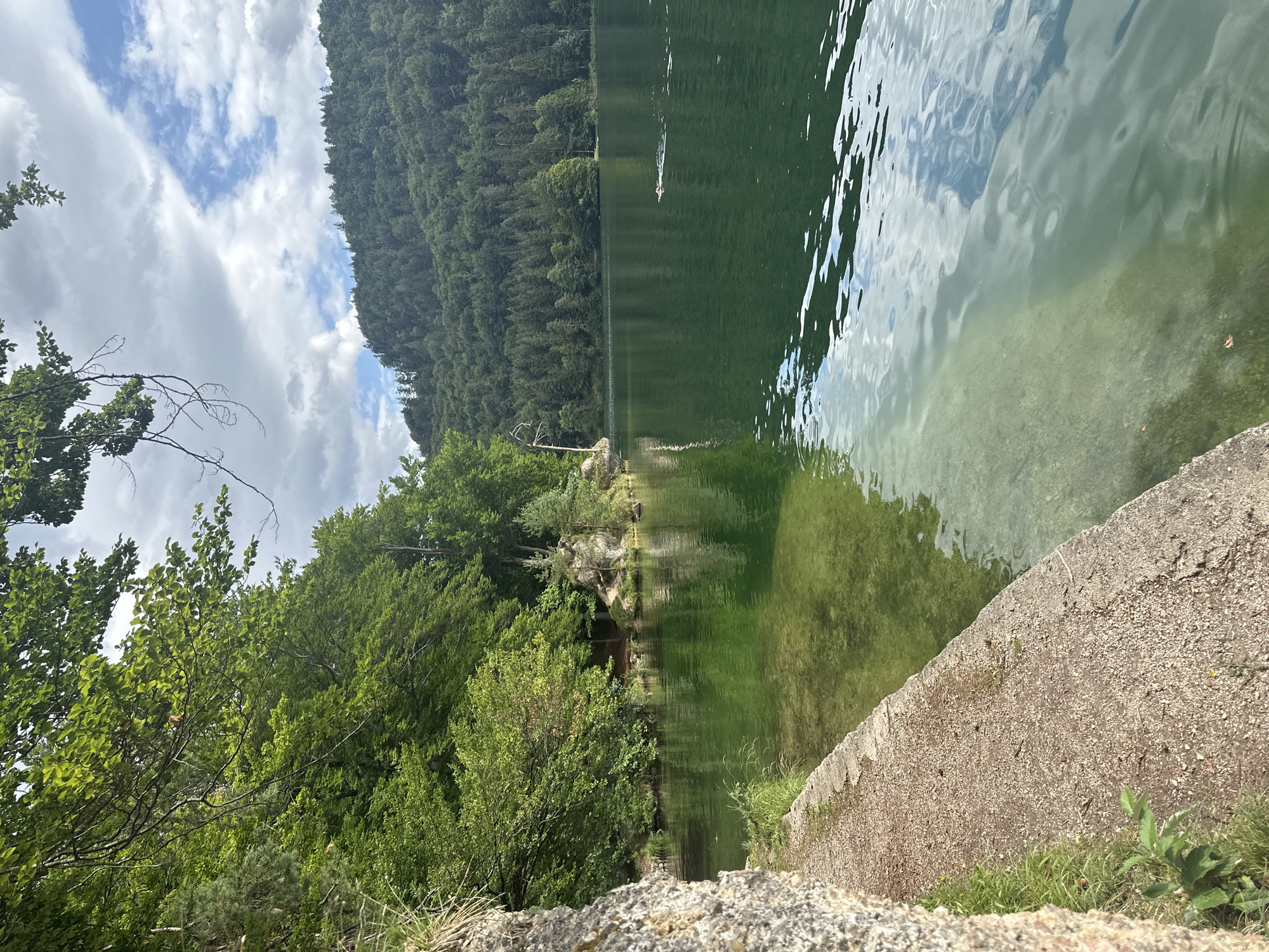 The image shows a serene lake scene, likely in the vicinity of Kufstein, Austria, given the lush forested mountains that surround the water. The foreground features a weathered stone embankment and a gravel path leading to the clear, green water. The lake is calm, with reflections of the sky and surrounding trees shimmering on its surface. In the distance, a densely wooded mountain slope rises majestically, its peak partly obscured by dramatic, cloud-filled skies. A lone individual is visible in the lake, swimming towards the right of the frame. The overall impression is one of tranquility and natural beauty.