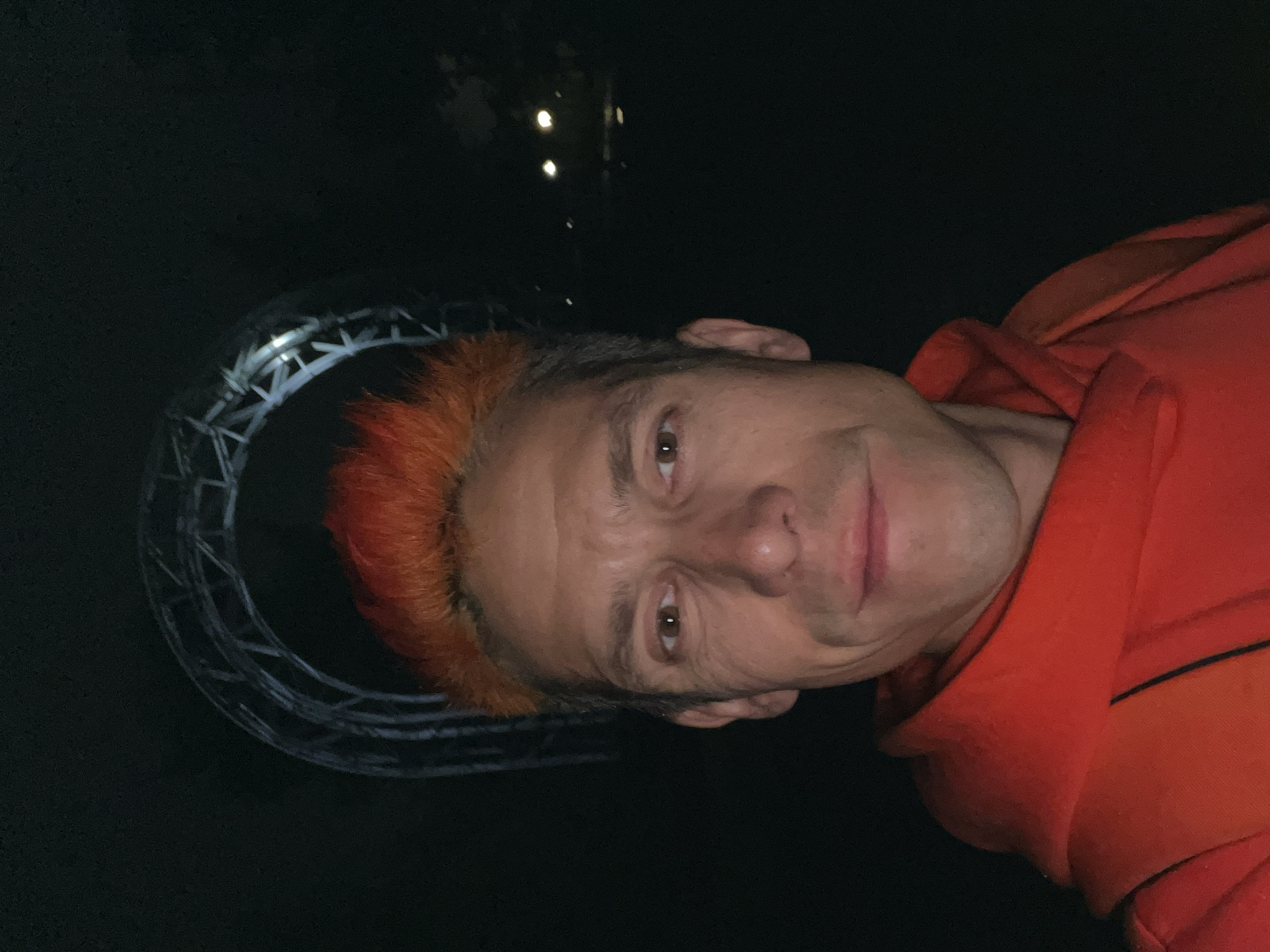 An adult male is captured in a close-up, head-and-shoulders portrait, appearing to be a selfie, set at night in Chaikovsky, Russia. He gazes directly into the camera with a slight, closed-mouth smile. His hair is short, spiky, and dyed a vivid orange. He wears an orange hooded sweatshirt. The background is predominantly dark. Above and behind his head, a large, metallic, arched truss structure is visible, consistent with a section of the Chaikovsky Bridge over the Kama River. The structure is dimly illuminated by ambient light, creating subtle reflections. Faint, blurred lights are discernible in the distant right background.