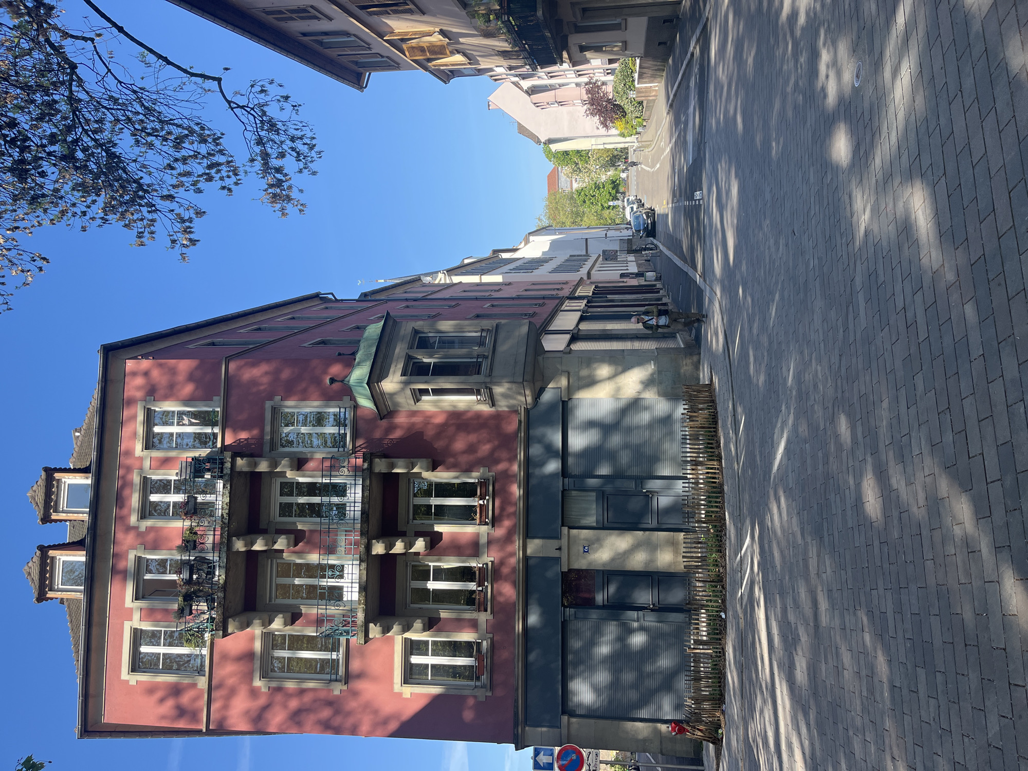 The image displays a sunlit street scene in Strasbourg, France, characterized by European urban architecture. The primary subject is a multi-story building with a deep red or pink facade on the left, featuring rows of white-framed windows and ornate wrought-iron balconies, some adorned with small plants. Stone lintels and sills frame the windows, and a distinctive green architectural detail accents the roofline on the top left.
To the right, a paved street, constructed from grey rectangular bricks, stretches into the distance. Long, distinct shadows from trees or overhead structures fall across the street and partially onto the ground-floor units of buildings, suggesting a clear, sunny morning or late afternoon. The sky above is a vibrant, cloudless blue, with the bare branches of a tree visible in the upper left corner, further contributing to the shadows.
Other multi-story buildings line both sides of the street, exhibiting a variety of pastel colors like light pink, white, and beige. Some of these buildings feature traditional window shutters. Further down the street, several cars are parked along the curb. No people are visible in the scene.
Notable details include a low wooden picket fence along the sidewalk in the mid-ground. On a grey building facade on the right side of the street, the number "9" is clearly visible. Near the bottom right, a circular blue sign with a white arrow indicates a one-way street, and another red-bordered circular sign with a diagonal line suggests a "no parking" zone. The scene conveys a tranquil, everyday atmosphere of a historic European city street.