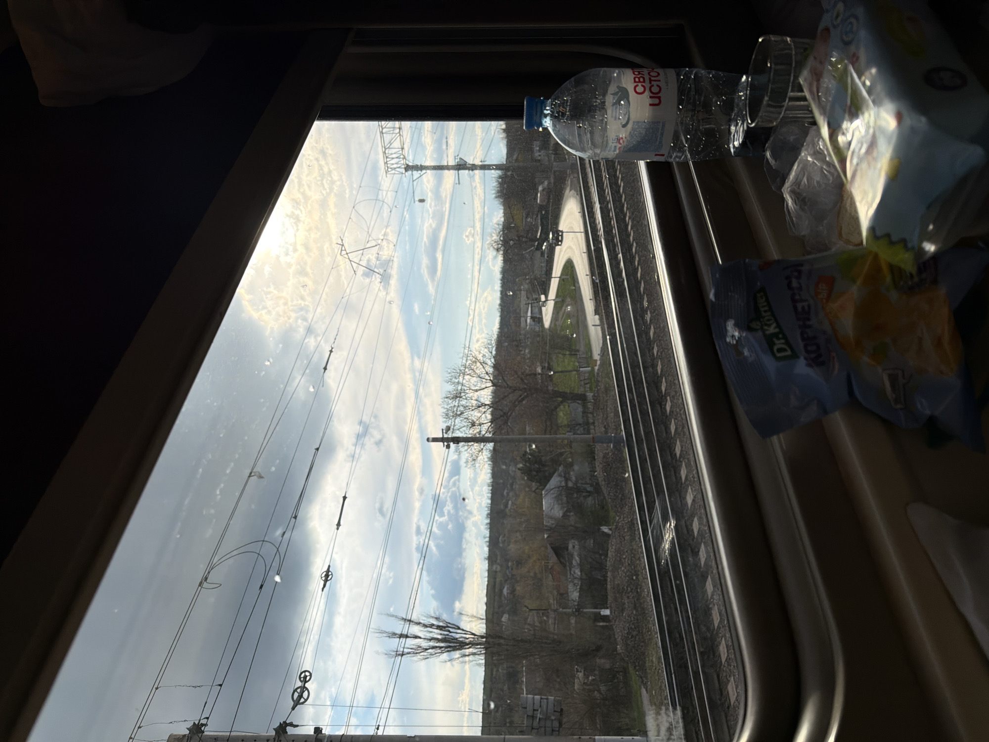 The image, apparently taken from inside a moving train, shows an outside view through a window. Beside the window on the right are a bottle of "Svyatoy Istok" water, and a bag of Dr. Korner chips. The view outside is of a rural landscape including railway tracks, transmission towers, trees, buildings, and roads. The sky is partly cloudy. The train is traveling through or near the city of Krasnyy Sulin, Russia, as evidenced by the brand of chips.