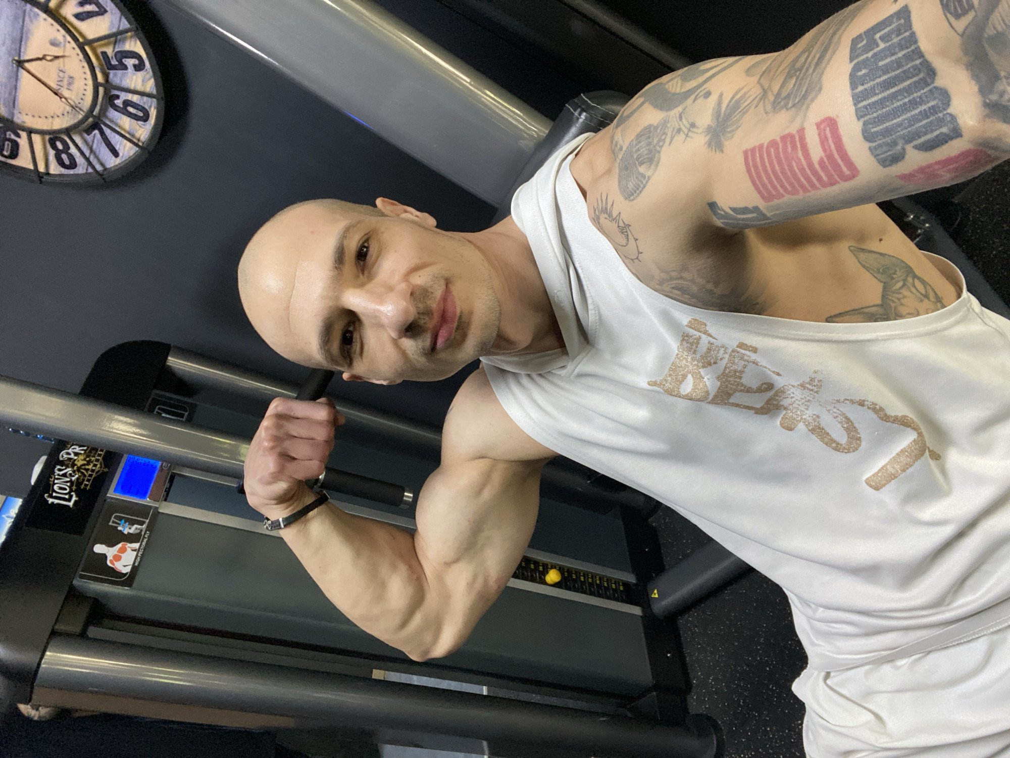 A bald, fair-skinned man is posing in what appears to be a gym. He's wearing a white hooded tank top with the word "Beast" printed on it and white shorts. He is flexing his right arm, which is heavily tattooed, as is his left arm, also tattooed. He wears a bracelet on his left wrist. In the background, a gym machine labeled "Lion's PR" can be seen, along with a clock on the wall, indicating the time is around 6:56. The location is likely in Pleven, Bulgaria.