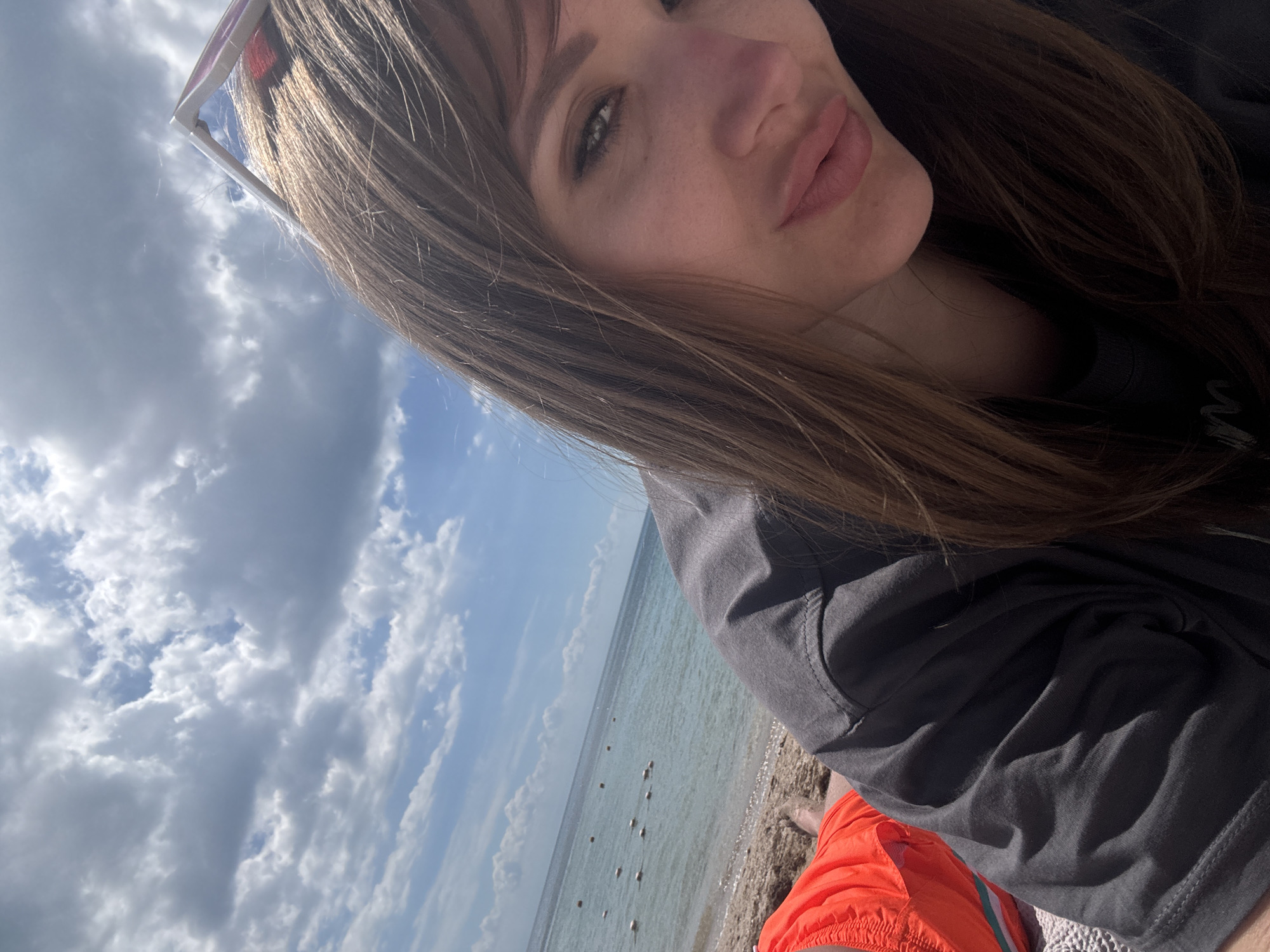 A woman with brown hair is seen in a selfie-style shot against the backdrop of a cloudy sky and a body of water. She is wearing sunglasses and a dark-colored shirt or jacket. The background features the sea with floats in the water. The beach appears to be sandy. The presence of the sea and beach suggests a coastal environment, likely in Yevpatoriya, Ukraine.