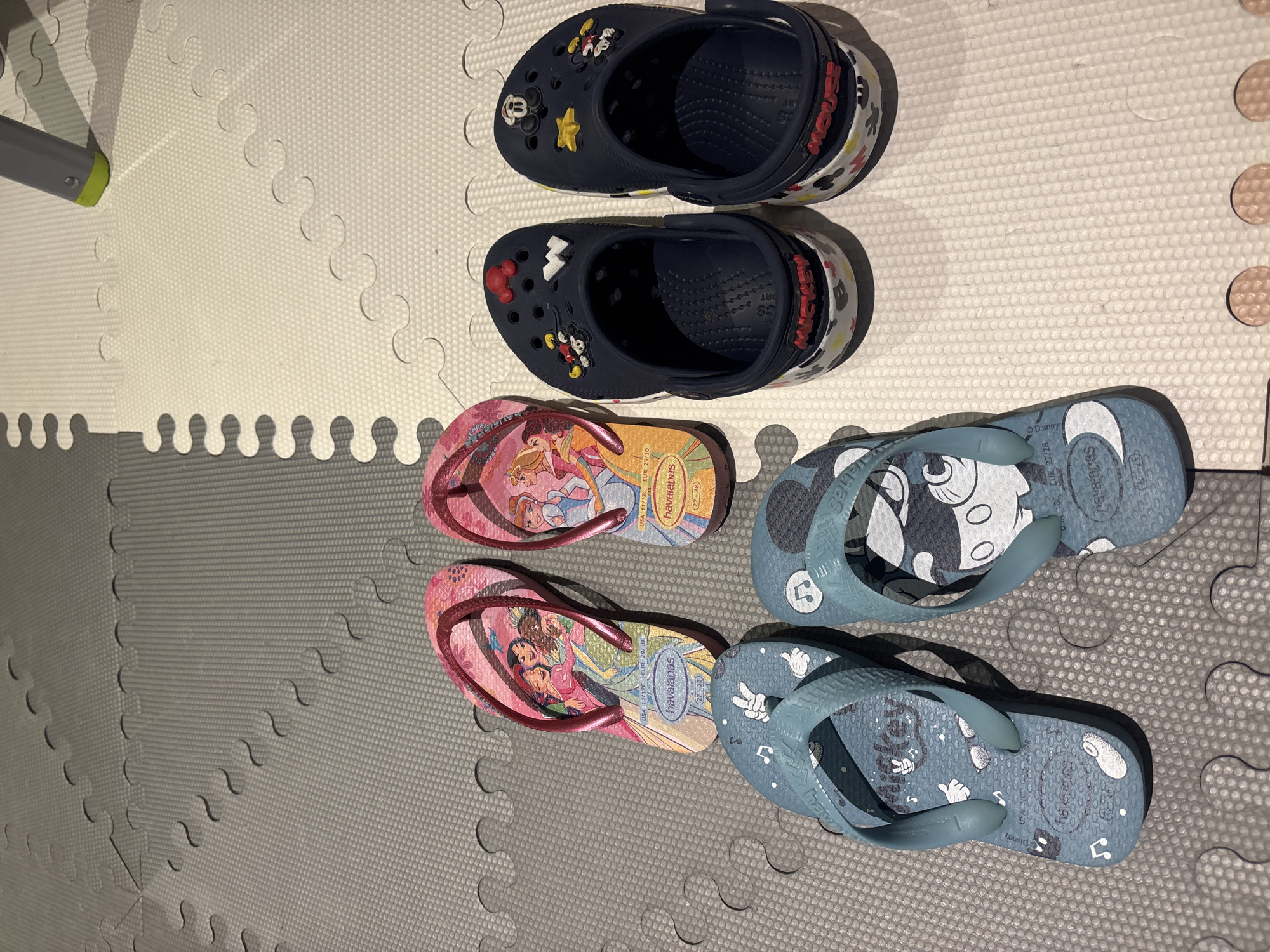 The image shows a collection of children's footwear arranged on a grey and white interlocking foam mat. On the left, there are two pairs of light blue Havaianas flip-flops featuring Mickey Mouse designs and musical notes. To the right of the flip-flops are two pairs of navy blue Crocs, also decorated with Mickey Mouse and other Disney-themed charms. In the background, two pairs of pink Havaianas flip-flops with princess designs are visible. The floor mat is placed on a neutral-colored surface. There are no people visible in the image. The scene is indoors, likely in a residential setting. There is no information in the image that suggests a location in Dubai, United Arab Emirates.