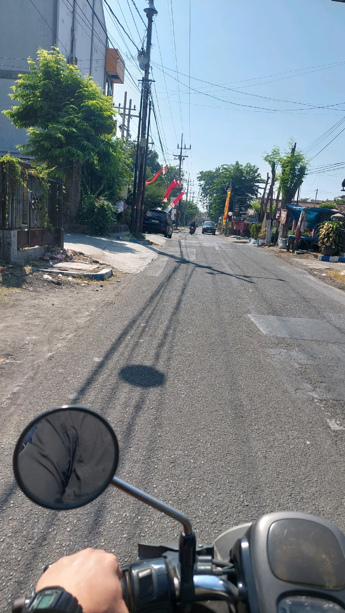 This is a first-person perspective image taken from a motorcycle on a paved road in Sidoarjo, Indonesia. The road appears to be a residential street with a moderate amount of traffic. The sky is clear and blue. On the left side of the road, there are houses with fences and lush green trees. Several red and white Indonesian flags are displayed, indicating a possible national celebration. A black SUV is parked on the left side of the road, and a few motorcycles are visible in the distance. The road surface shows some signs of wear and tear, with patches of asphalt repair. The perspective suggests the motorcycle is moving forward. The image captures a slice of everyday life on a sunny day in a suburban area of Sidoarjo.