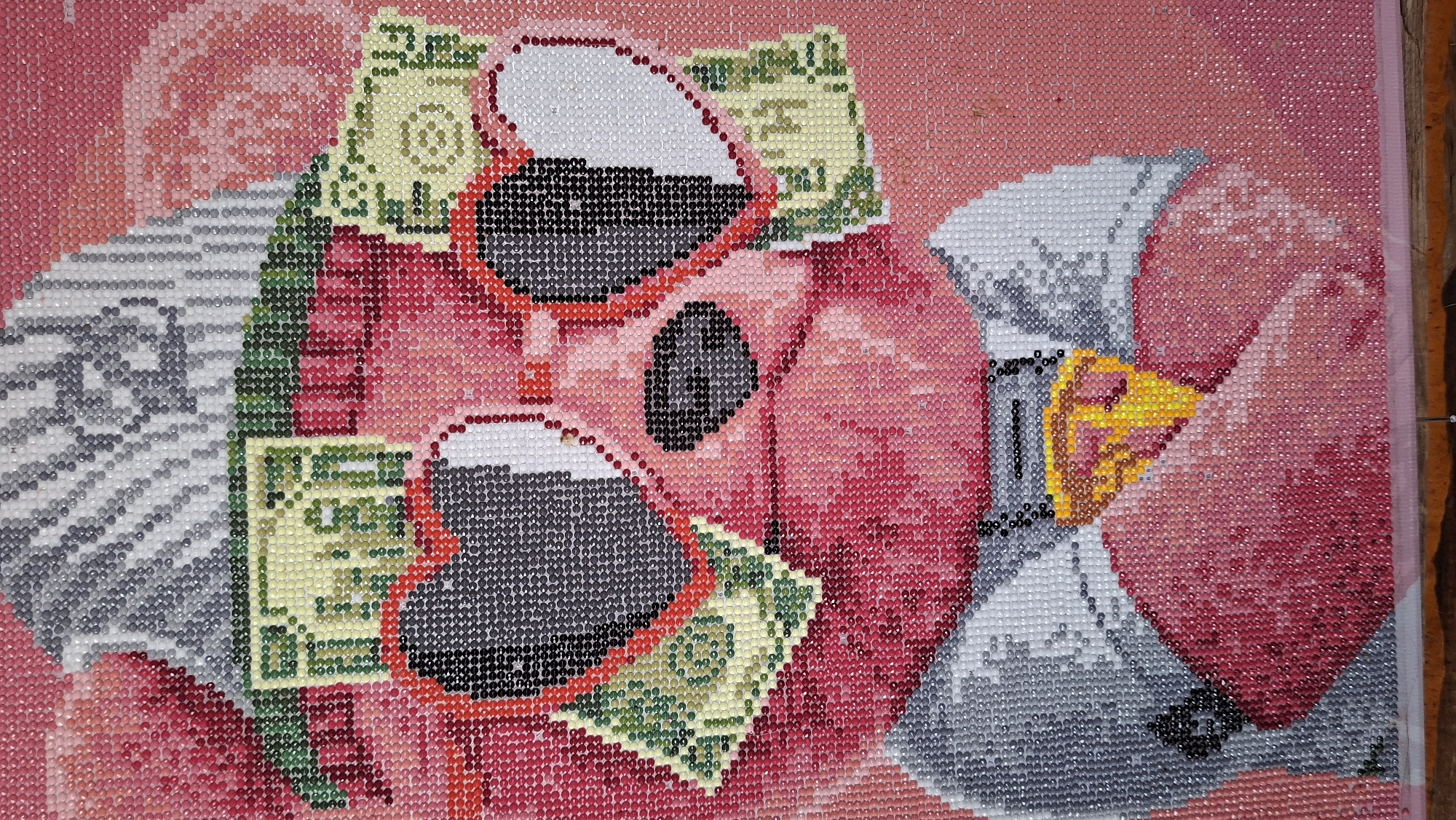 The image is a close-up of a diamond painting. The subject is a flamingo wearing heart-shaped sunglasses and holding a dollar bill in its beak. Additional dollar bills are placed around the flamingo's head. The background is a mottled pink. The diamonds used to create the image are small and uniform in size, giving the painting a pixelated appearance. There is no indication of the location Большой Букор, Russia, within the image.