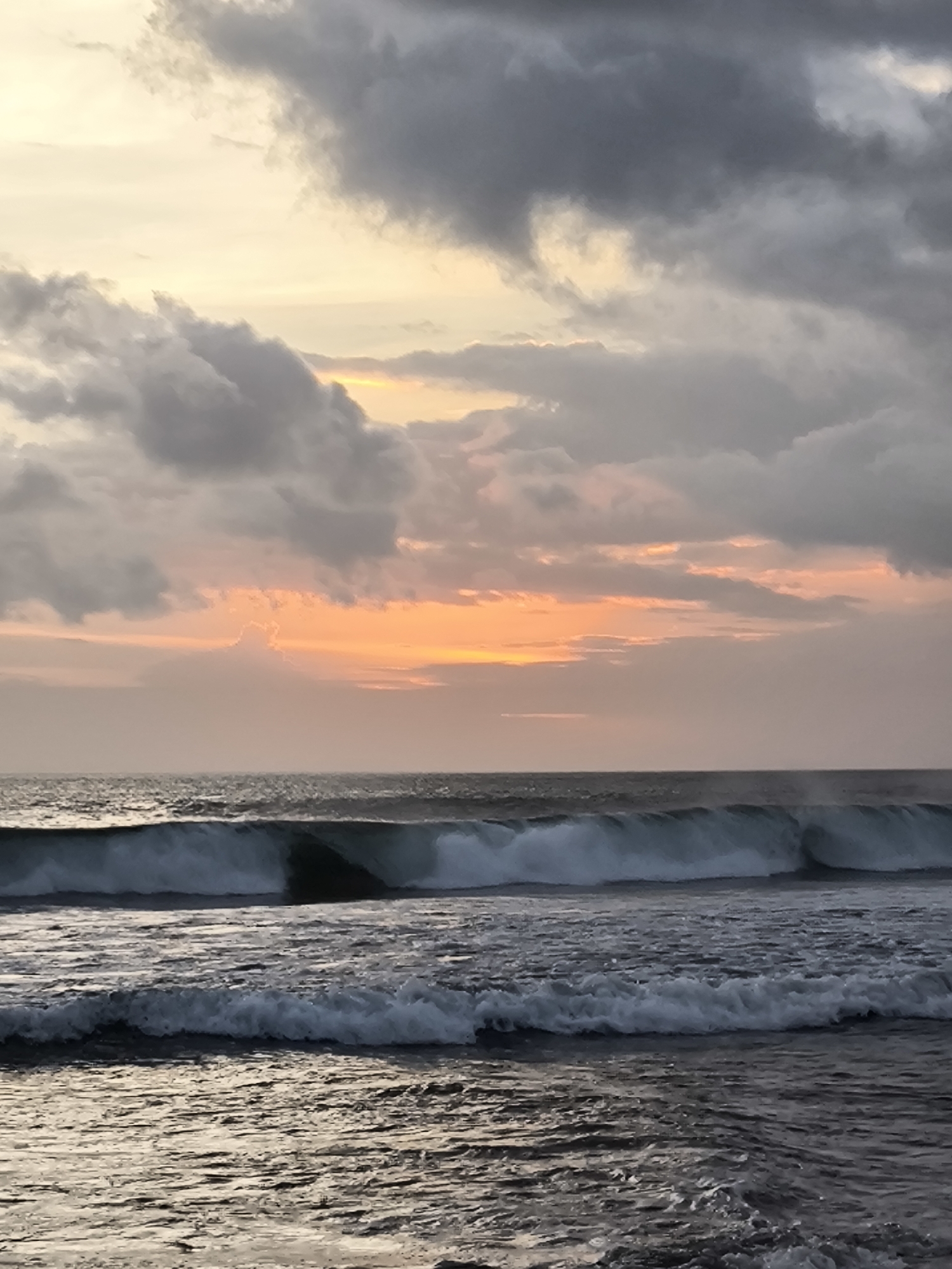 The image depicts a seascape at sunset in North Kuta, Indonesia. The sky is filled with dark, grey clouds with patches of orange and yellow light filtering through near the horizon. The ocean features rows of waves with white foam crests, rolling towards the foreground. The water in the foreground is dark and rippled. There are no people visible in the image. The scene is bathed in the soft light of the setting sun.