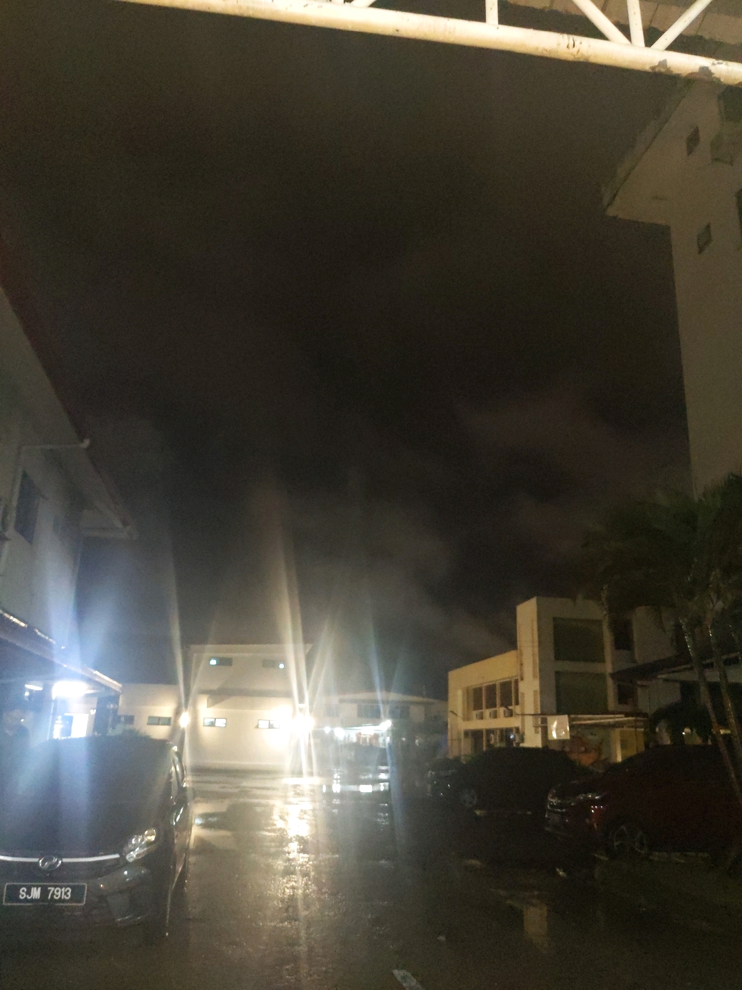 The image captures a nighttime scene on a wet, paved surface, likely a parking area, with several vehicles present. A black car with the license plate "SJM 7913" is prominently positioned in the foreground on the left. Further back, other vehicles are parked, their shapes obscured by darkness and reflections on the wet ground. Buildings with illuminated windows are visible in the background, suggesting an urban or commercial area. The sky is dark and appears cloudy, with bright lights emanating from the buildings and streetlights creating lens flares and illuminating the mist or fog. The overall atmosphere is damp and dimly lit. The scene is located in Kolombong, Malaysia.