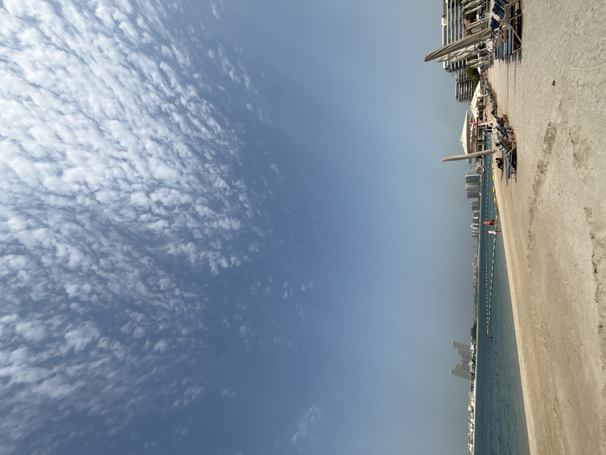 This is a vertical, eye-level, outdoor, daytime photograph of a beach scene in Dubai, United Arab Emirates. The sky occupies the upper two-thirds of the frame, with a clear blue expanse populated by scattered, wispy white clouds. The lower third of the image features a sandy beach, the turquoise ocean, and a distant cityscape.
The beach is lined with rows of lounge chairs and umbrellas, many of which are unoccupied. A few people are visible on the beach and in the water. On the sand, two individuals are sitting in lounge chairs, and two other figures are standing near the water's edge. In the water, a couple is visible.
The ocean is calm and appears shallow near the shore, marked by a yellow floating barrier. In the background, a cluster of modern high-rise buildings is visible along the coastline. The architecture is contemporary, with angular designs and reflective glass facades. The overall impression is one of a well-maintained resort or public beach area. The lighting suggests a bright, sunny day.