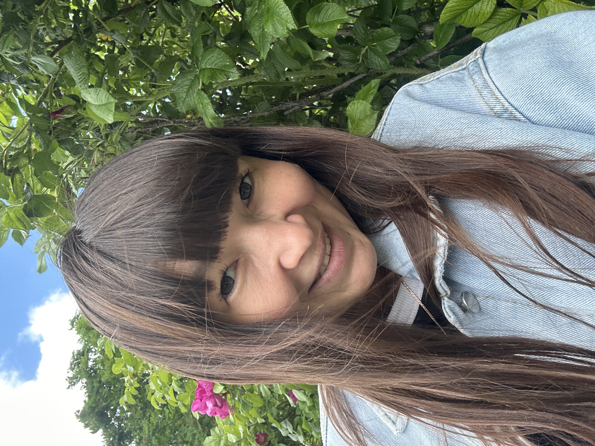 In a close-up outdoor selfie, a woman with long, brown hair and bangs smiles at the camera. She is wearing a light blue denim jacket over a dark top, and a silver necklace is visible. Behind her, a dense bush with vibrant green leaves and a pink rose is present. The sky above is blue with scattered white clouds. The scene appears to be in a natural outdoor setting, possibly a park or garden, given the foliage. The photograph was taken in Chaikovsky, Russia.
