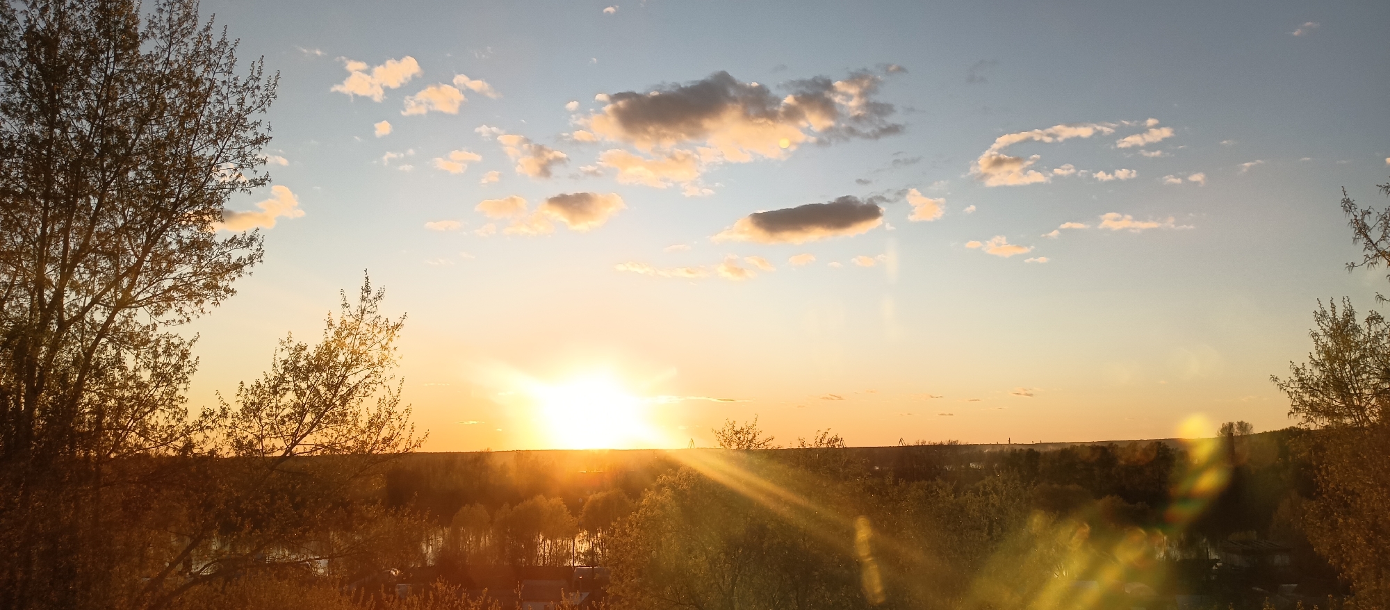 The image shows a sunset scene. The sun is setting on the horizon, creating a bright, golden glow. The sky is a mix of blue and orange hues. There are scattered clouds in the sky, some with golden edges from the sunlight. The scene is viewed from an elevated position, possibly a hill or a high building, looking over a landscape with trees and some visible buildings. Some of the trees are silhouetted against the bright sky, while others have a golden glow. The location of the scene is unknown.