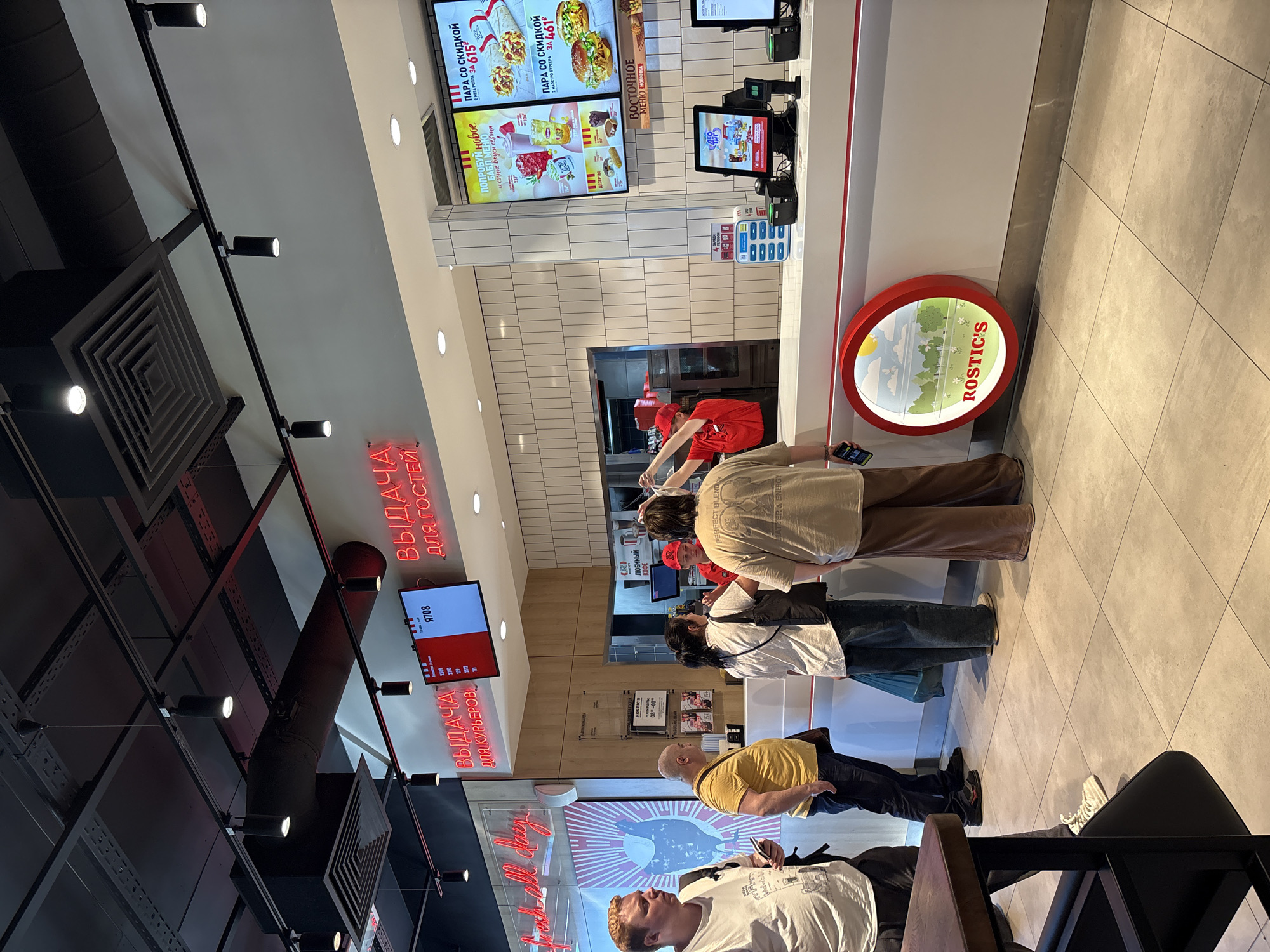 A group of people are standing in line at a fast-food restaurant. The restaurant appears to be a Rostic's, based on the logo visible on the counter. Several digital menu boards are displayed above the counter, showing various food items and prices. A digital display above the counter shows a ticket number. A staff member in a red uniform is visible behind the counter, interacting with customers. The interior of the restaurant is modern and clean, with white tiled walls and a white counter. The floor is tiled in a light color. The lighting appears to be a mix of track lighting and recessed lights. The overall scene suggests a typical busy day at a fast-food establishment.