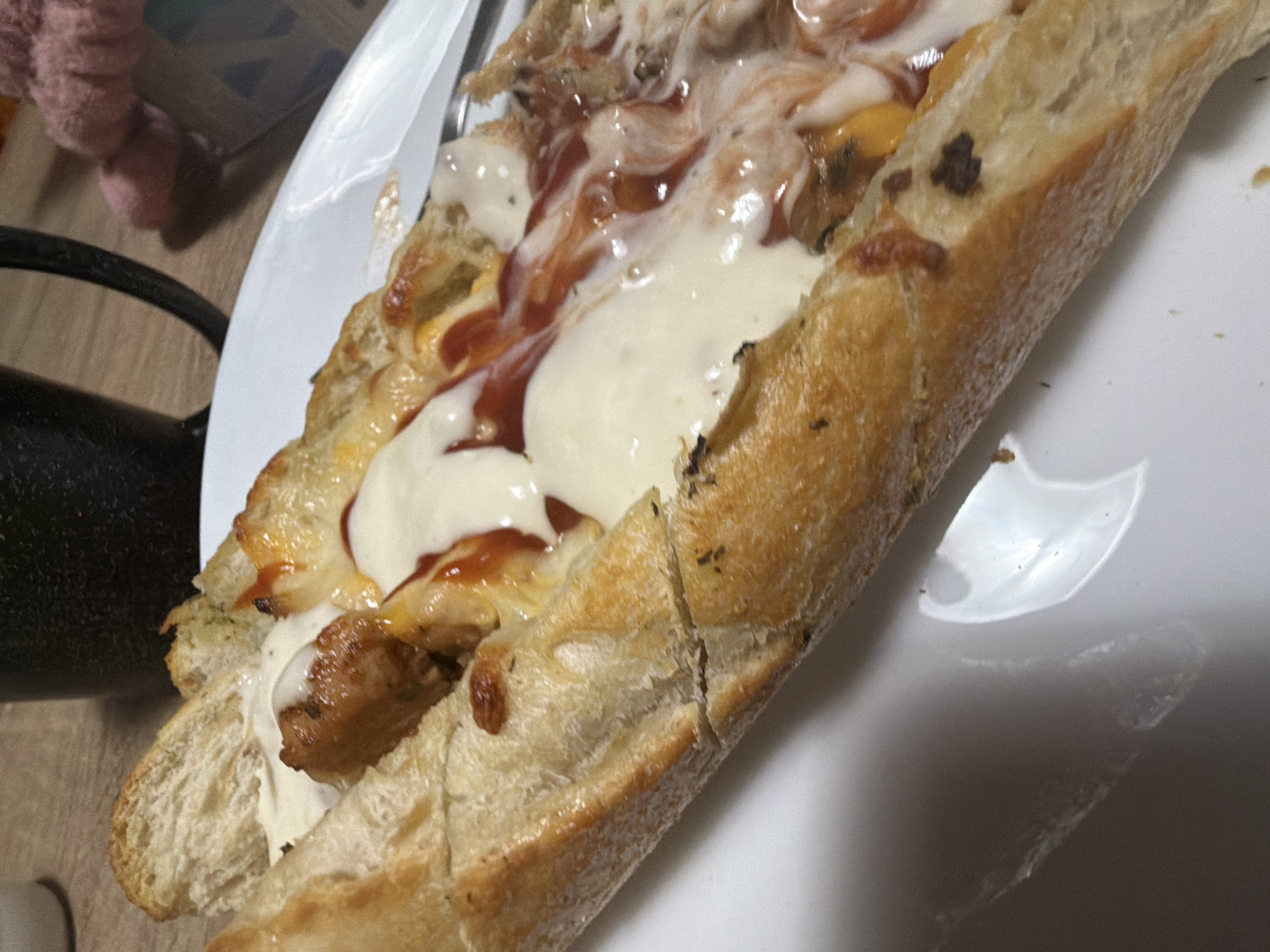 The image shows a close-up of a long sandwich served on a white plate. The sandwich appears to be made with a baguette-style bread that has been split lengthwise and filled with ingredients that include what look like meatballs or sausage, melted cheese, and a red sauce, possibly ketchup or marinara. The bread is golden brown and appears to have some herbs baked into it. The filling is generously layered and is partially visible spilling out of the bread. The lighting suggests an indoor setting, possibly a home or a restaurant. The background is blurred but shows a wooden surface, a dark mug or cup, and a pink fuzzy object. There is no discernible information in the image to suggest a specific location in Plovdiv, Bulgaria.