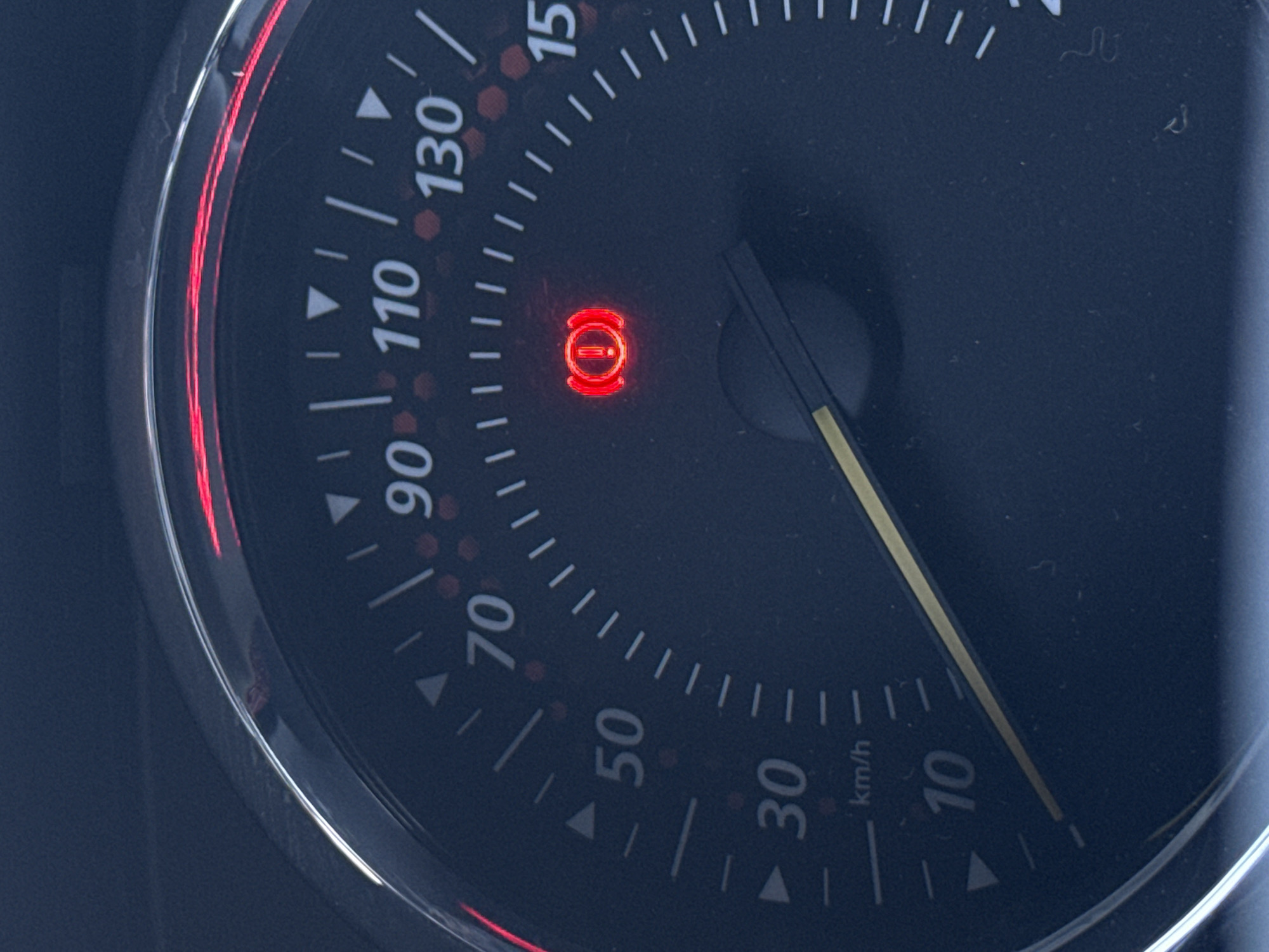 The image shows a portion of a car's dashboard. The speedometer is visible, with the needle pointing to approximately 6 km/h. A red warning light in the shape of a circle with a line through it (likely indicating a brake system issue) is illuminated. The scale of the speedometer goes up to 150 km/h. The image lacks details to establish a specific location, let alone Izhevsk, Russia.
