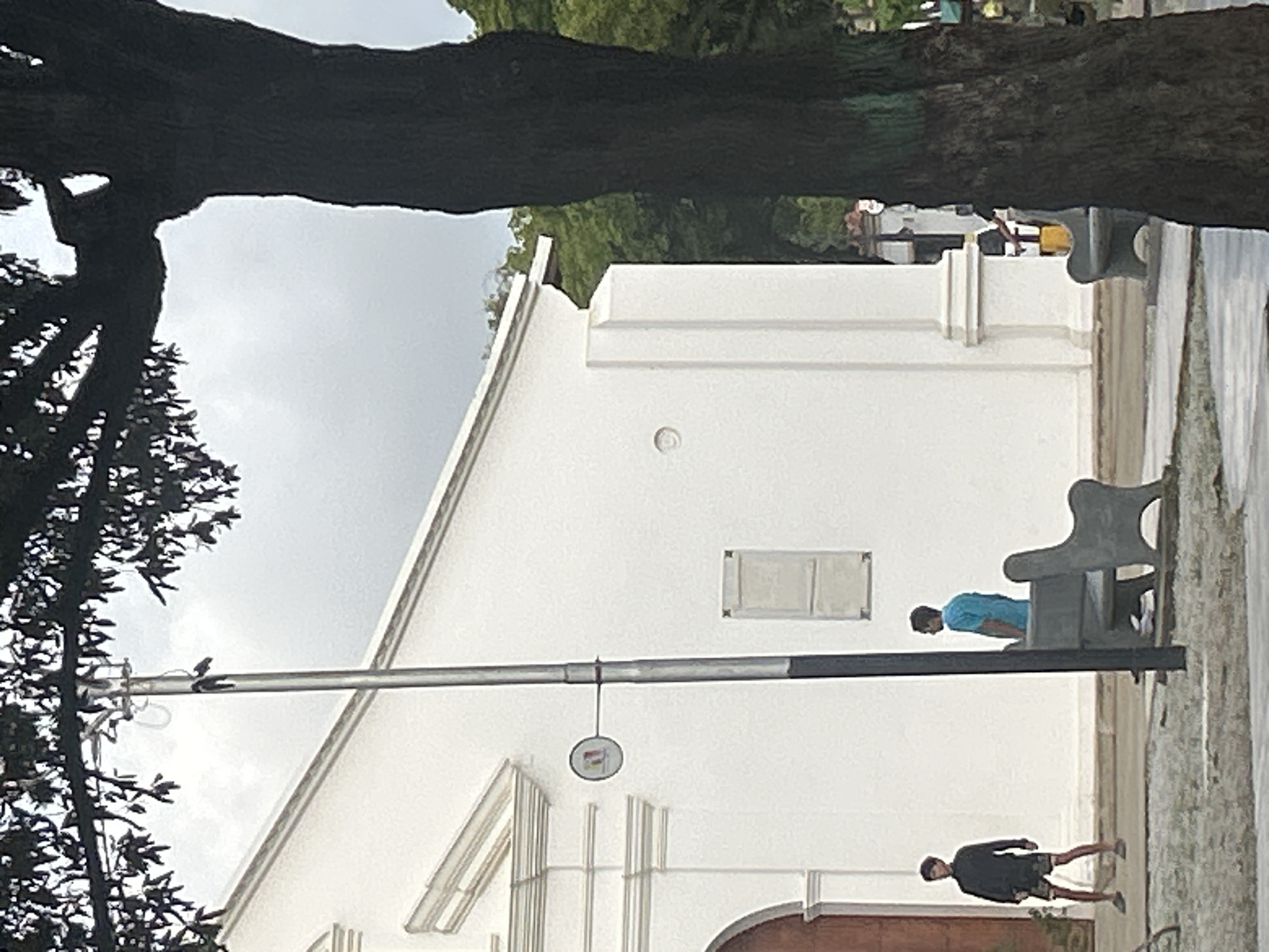 The image depicts a street scene in Santa Ana, Venezuela, with a white building resembling a church or public building as the backdrop. A large, dark tree trunk dominates the right side of the frame, obscuring a portion of the building and the sky. In the foreground, two men are visible. One man on the left wears a dark jacket and patterned shorts, standing near a large wooden door. Another man on the right wears a blue t-shirt and stands near a concrete bench. Another bench is visible to the far right. A lamppost stands prominently in the center of the image, with a circular sign attached to it. The ground in the foreground appears to be paved or covered with gravel. The sky is overcast, suggesting a cloudy day. The architectural style of the building suggests a colonial or traditional design.