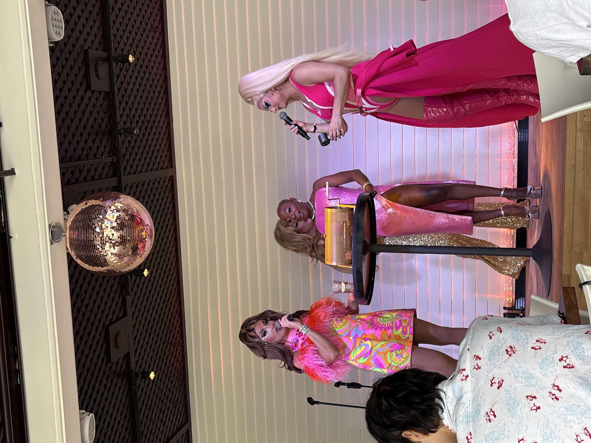 In Provincetown, Massachusetts, at an indoor venue, three drag queens are performing on a stage in front of a white, slatted wall. The queen on the left wears a vibrant, psychedelic-print dress with pink feather trim and holds a microphone. The queen in the middle, with a shaved head, wears a sparkly pink dress and is holding a golden lottery-style raffle drum. The queen on the right has long blonde hair and wears a bright pink outfit with a dramatic cape and thigh-high boots, also holding a microphone. A disco ball hangs from the ceiling above the stage. In the foreground, a person wearing a patterned shirt is seated, with their back to the camera, observing the performance. The lighting is dramatic, with pink and purple hues cast on the stage and wall.