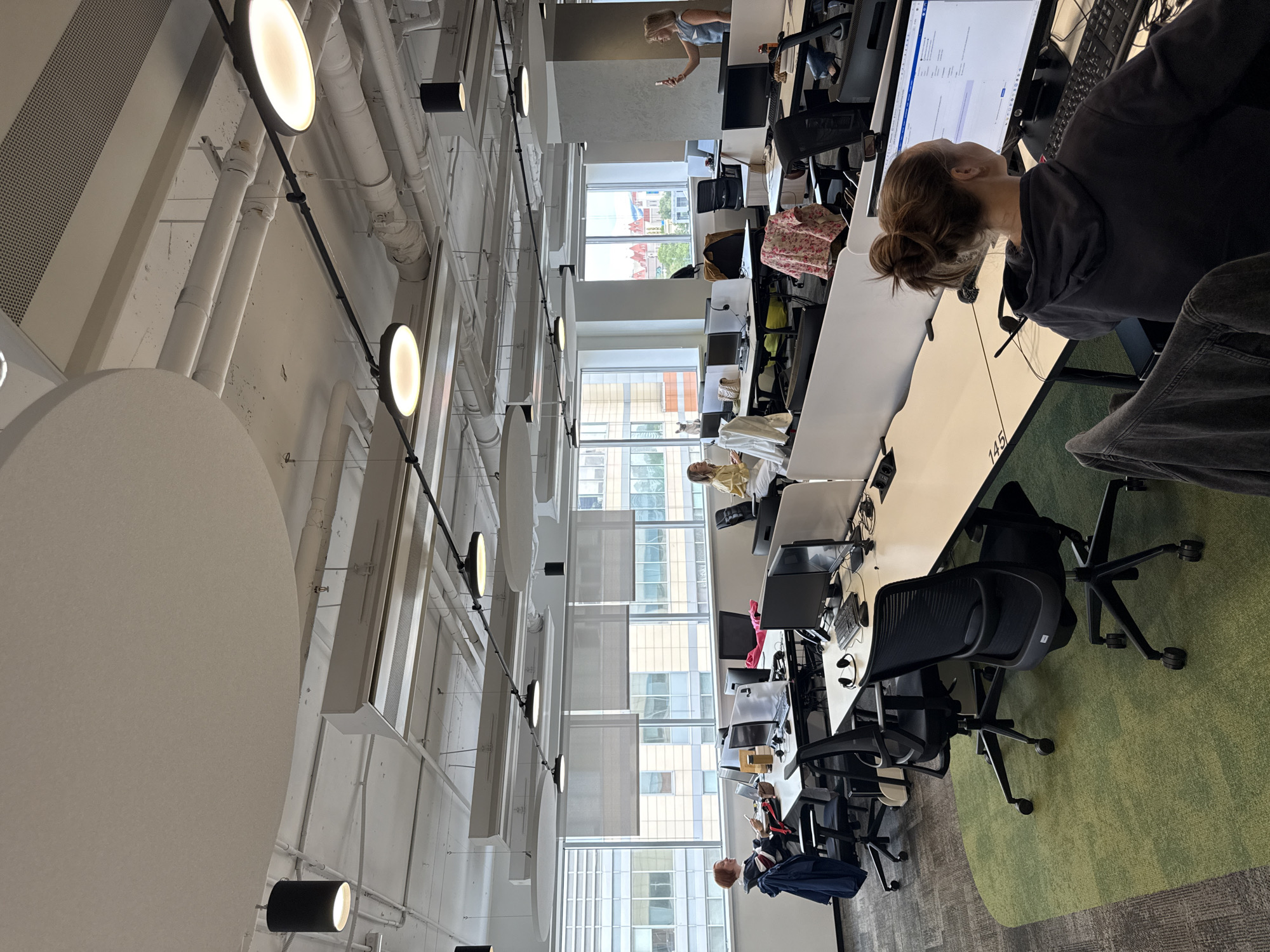 The image depicts an office space in a modern building, likely located in a city given the view from the windows. The interior is characterized by a long row of desks, each equipped with a computer and chair, indicating a workplace. Employees are scattered throughout the space, some sitting and working at their desks, while others are standing and possibly interacting or engaged in activities. The lighting is provided by a series of round light fixtures suspended from the ceiling, and the presence of windows allows for natural light to enter the room. The overall environment is functional and organized, suggesting a professional setting. The floor appears to be carpeted with a green carpet and gray carpet.