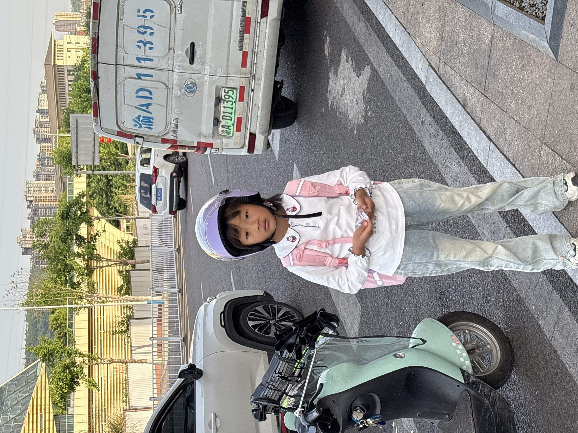 A young Asian girl, wearing a lavender helmet, a white long-sleeved shirt with strawberry accents, light-wash jeans, a pink backpack, and white sneakers, stands on the sidewalk next to a mint green scooter parked adjacent to a white car. The girl is holding something in her hands and looking at the camera. Behind her, a white van with the license plate 渝A D11395 is parked on the street. The scene appears to be in an urban setting, potentially in Jiangjin, China, as indicated by the "渝A" license plate prefix which denotes Chongqing. The buildings and trees in the background suggest a dense residential area.