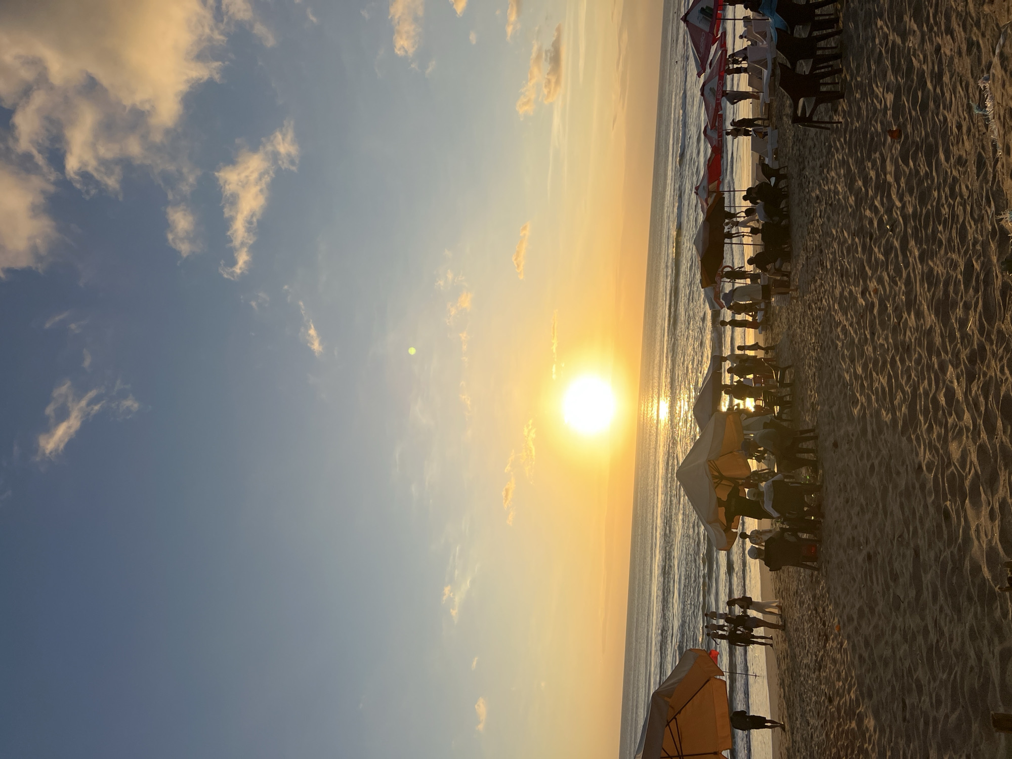This is a photograph of a crowded beach at sunset in Kuta, Indonesia. The sun is low on the horizon, casting a warm, golden glow across the scene. The ocean is calm with gentle waves, and the sky is a gradient of blue and orange, with scattered clouds tinged with yellow.
In the foreground, there are several beach umbrellas and chairs set up on the sandy beach. People are scattered throughout the scene, some sitting under the umbrellas, others walking along the shore, and a few are visible in the water. The individuals appear to be tourists and locals enjoying the sunset. The beach is busy with activity, suggesting a popular tourist destination.
The overall atmosphere is one of relaxation and enjoyment, with the natural beauty of the sunset enhancing the scene.