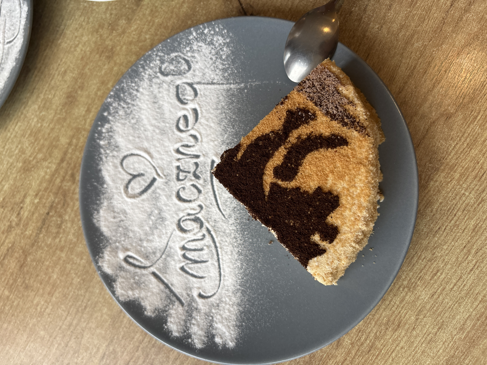 A slice of tiramisu sits on a gray plate dusted with powdered sugar, forming the word "macznego" and a heart. A spoon rests on the right side of the plate. The scene is set on a wooden table. The image was taken in Hajnówka, Poland.