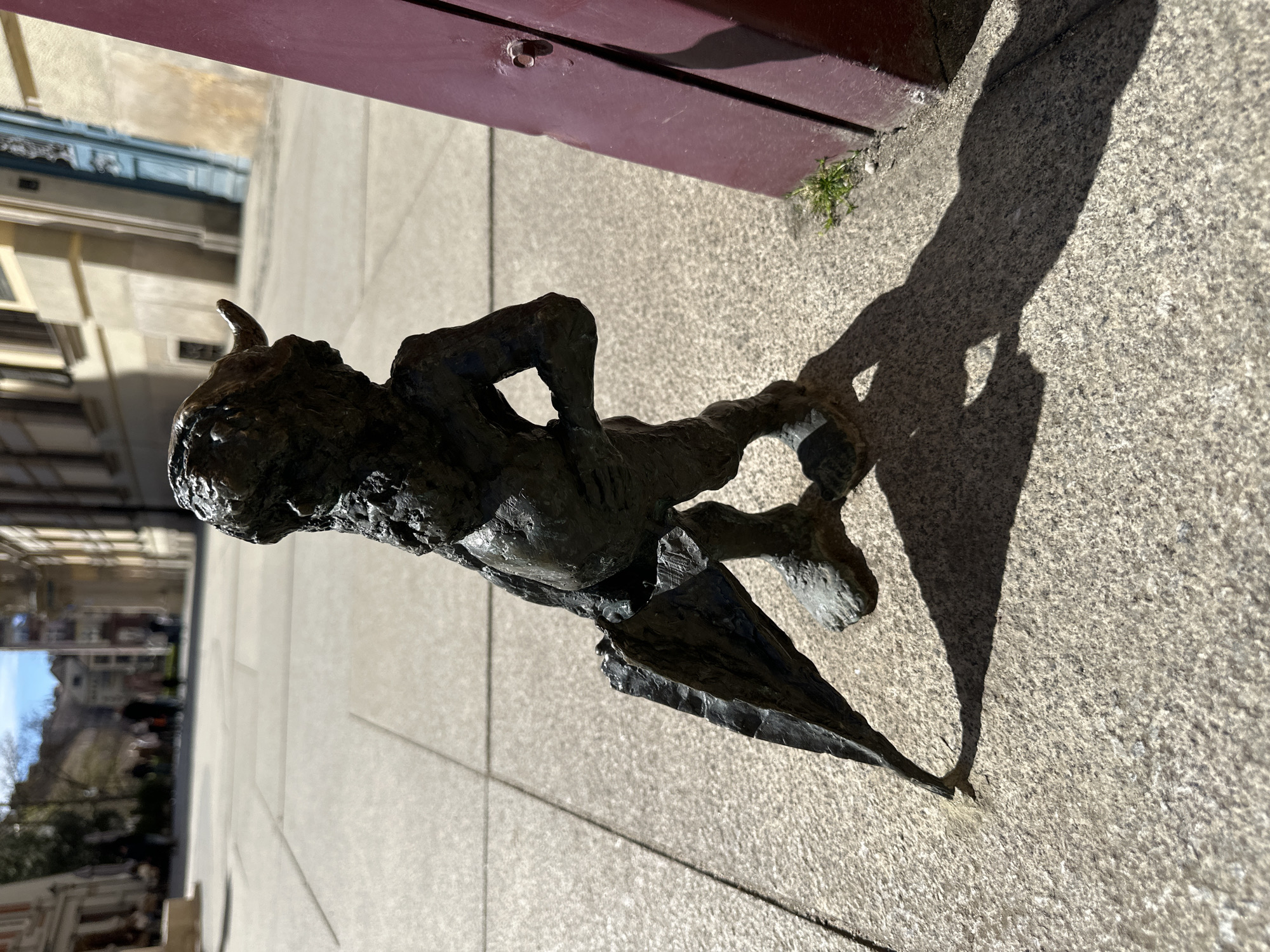 A bronze dwarf statue, one of many scattered throughout the city of Wrocław, Poland, stands on a paved surface next to a building. The statue has a grotesque appearance, with a horn-like projection from its head. It is positioned near a reddish-brown door or wall. The statue casts a shadow on the pavement. The background shows buildings across the street.