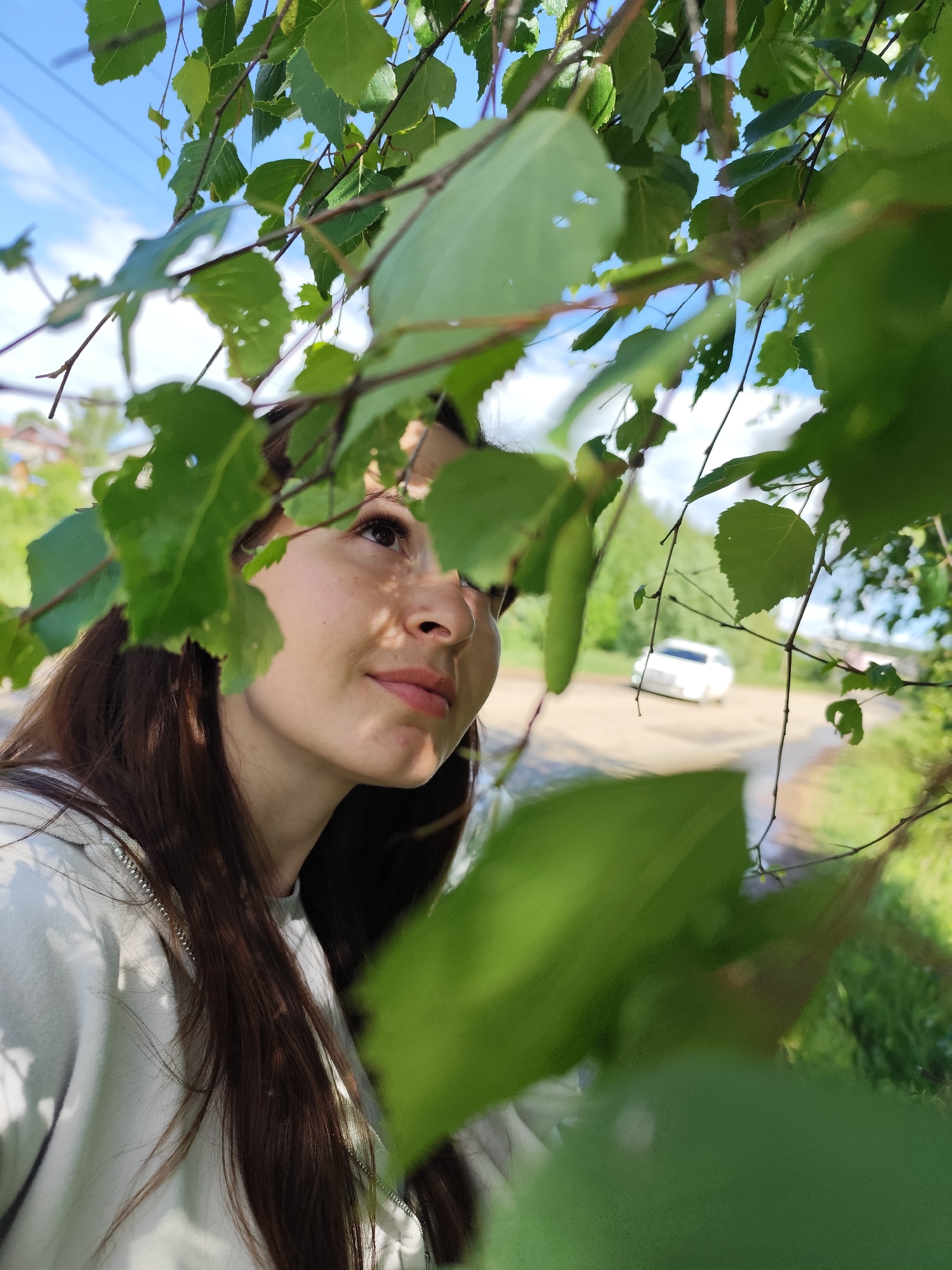 A woman with long brown hair is looking up and to the right, partially obscured by green leaves. She is wearing a light-colored top with a visible zipper. The background shows a street with a white car and greenery. The location is Фоки, Russia, based on the provided information. The sky is visible between the leaves.