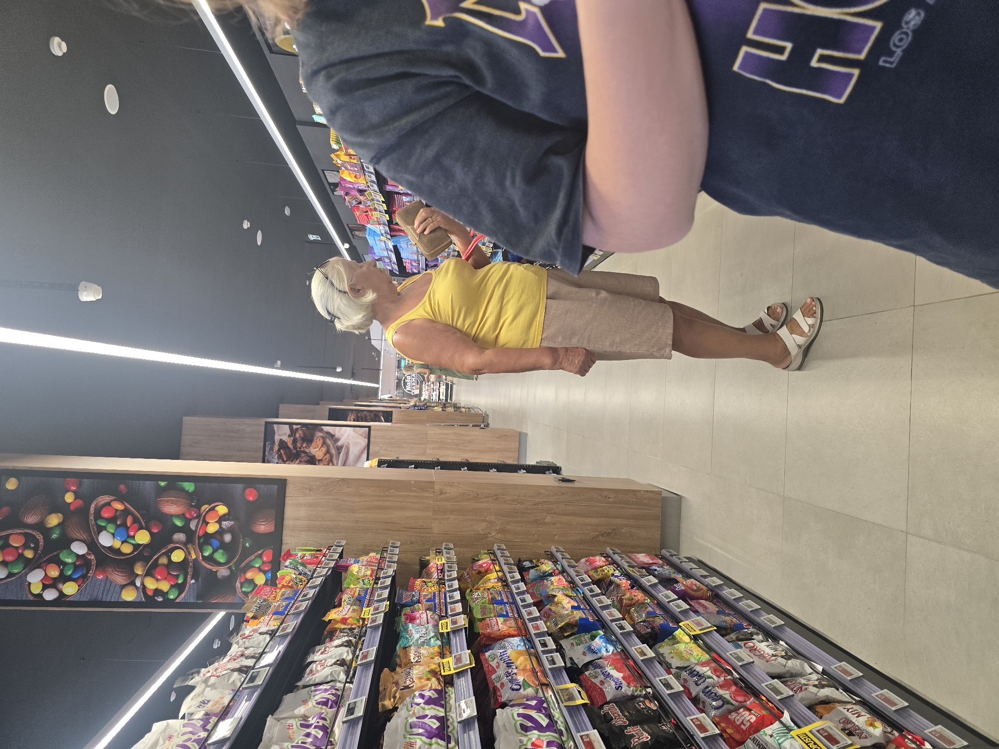 An exterior shot captures a woman with short blonde hair, wearing a yellow tank top and beige shorts, standing in an aisle of a supermarket. She is holding a wallet and looking at a shelf stocked with snacks. She is wearing white sandals. In the foreground, another person's shoulder and arm are visible, wearing a dark blue t-shirt with a purple and white logo. The supermarket aisle is well-lit with fluorescent lights and features shelves displaying various packaged food items. A large poster featuring colorful Easter eggs in chocolate shells is visible on a pillar to the left. The floor is tiled with large, light-colored squares. The environment appears to be a modern retail space, likely a grocery store in Podgorica, Montenegro, given the context.