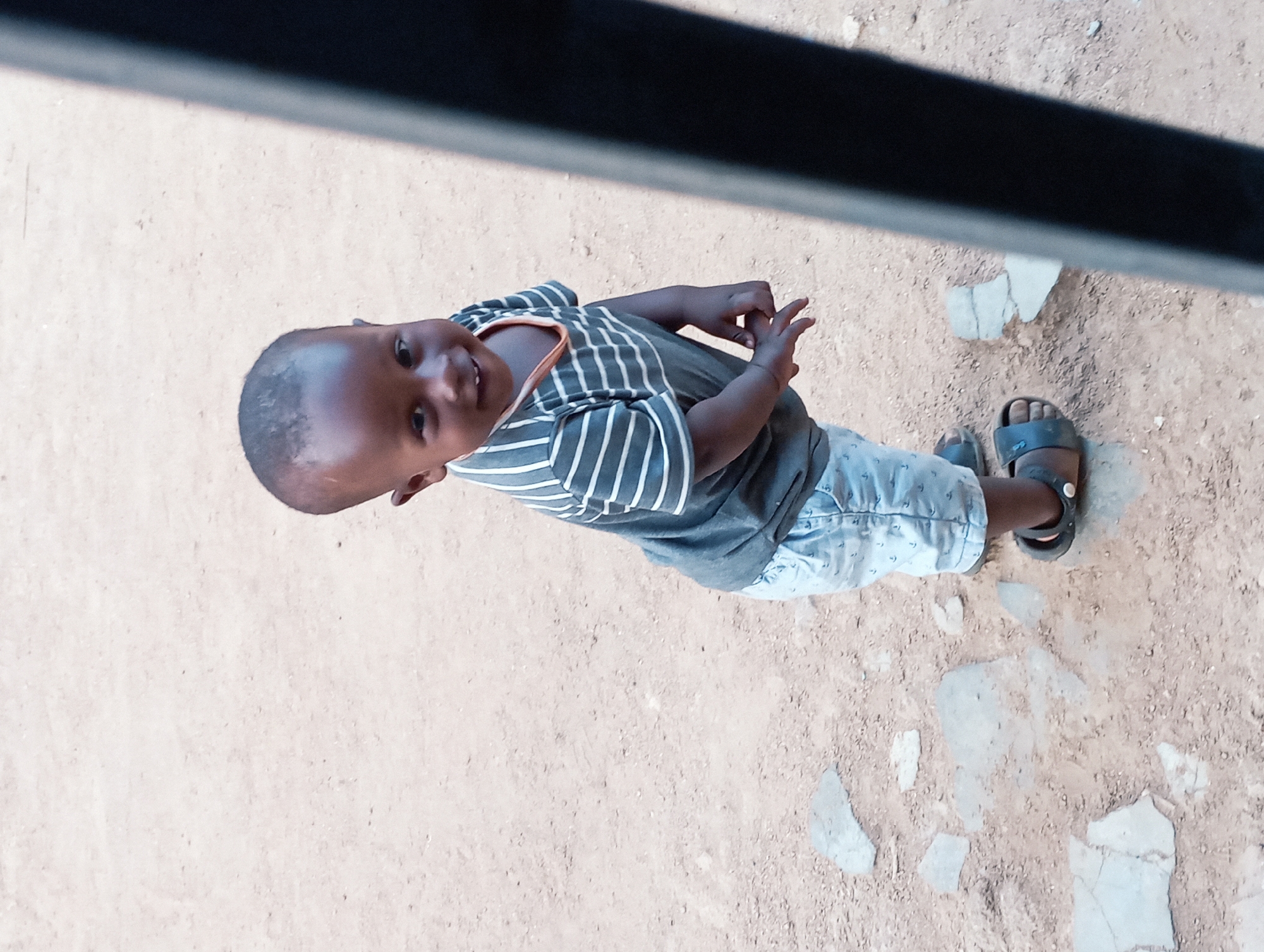 A child with dark skin and a shaved head stands on a dirt surface littered with small, light-colored rocks. The child wears a gray and white striped shirt, light blue patterned shorts, and dark sandals. He looks up and smiles, holding his hands up slightly. The child is outside, possibly in the city of Jos, Nigeria, indicated by the style of clothing and the environment. Overhead is a blurry dark gray or black structure, potentially part of a building or roof. The dirt surface appears dry and somewhat compacted.